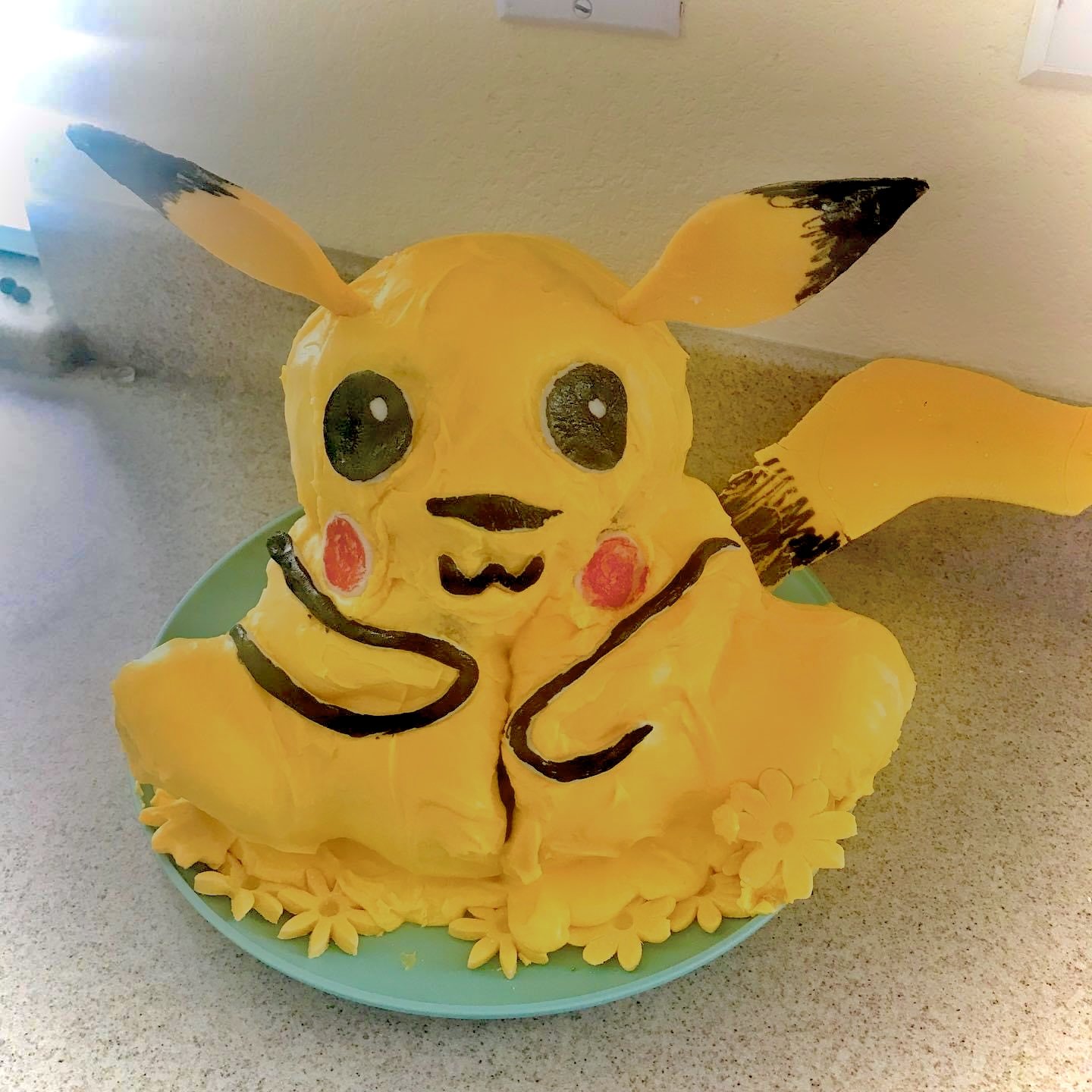 A cake decorated as Pikachu, a yellow Pokémon character with large black eyes, pink cheeks, and black-tipped ears, made with yellow frosting and detailed with black icing, sitting on a green plate surrounded by yellow flower decorations.