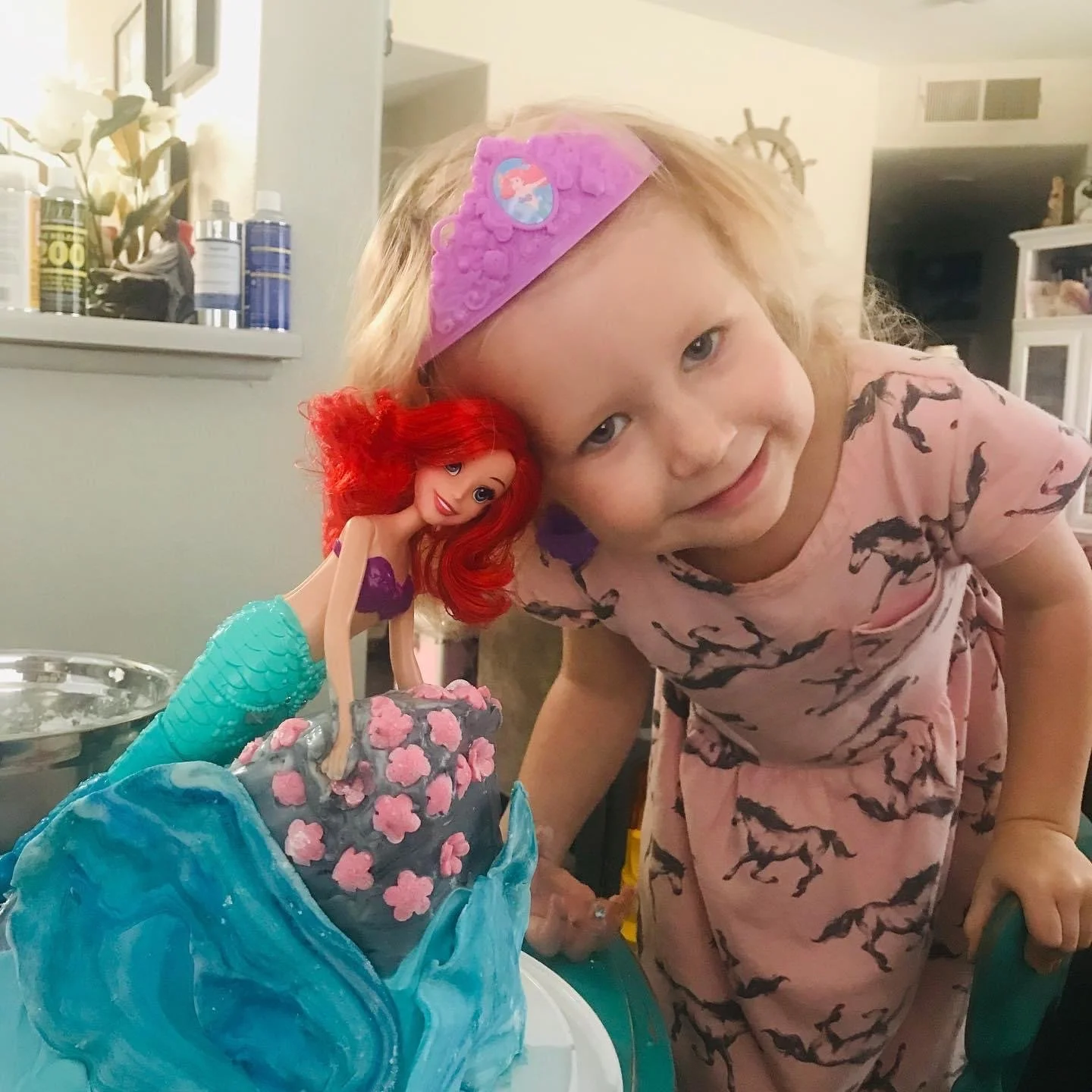 A young girl in pink pajamas with black horse patterns leaning over a Disney Ariel mermaid birthday cake decorated with pink flowers and blue ocean waves, with a small Ariel figure on top, during a birthday celebration.