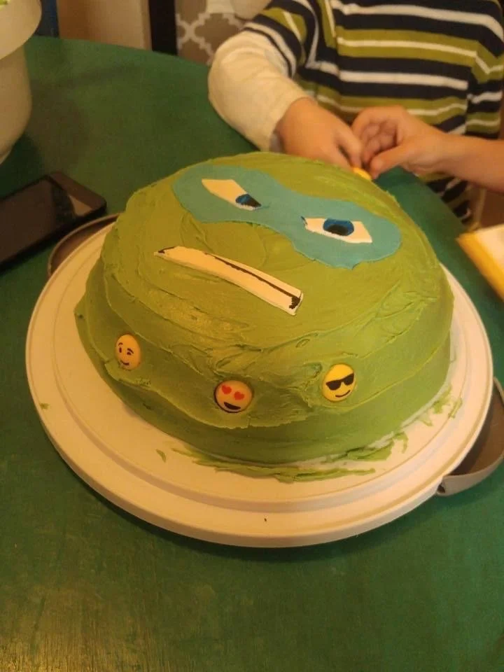 Green birthday cake decorated with a cartoon face, blue eyes, and emojis around the base, on a white cake stand on a green table. Two people are sitting at the table, one wearing a striped shirt.