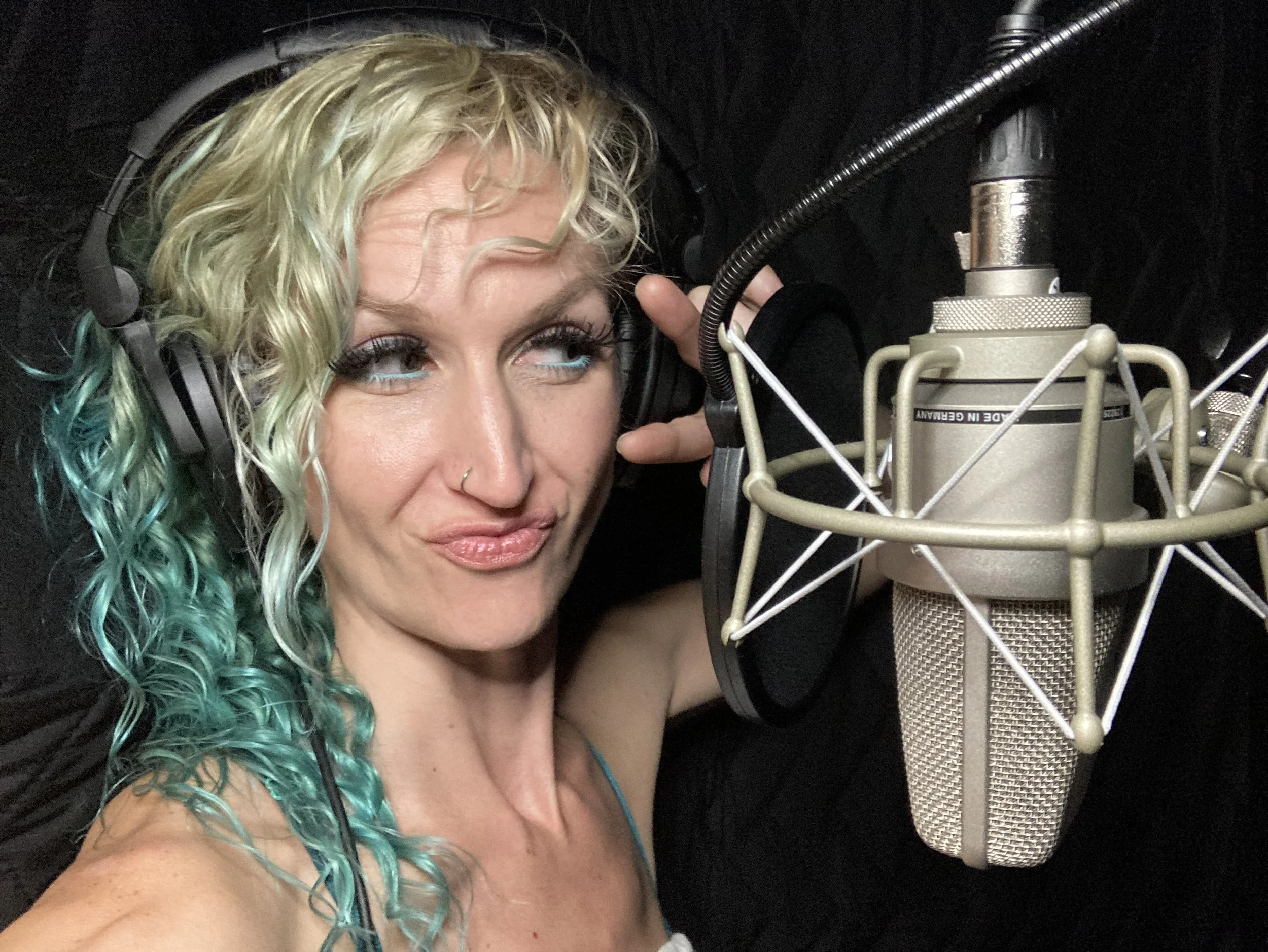 A woman in a recording studio wearing headphones, with her hand near her ear, standing close to a large condenser microphone.