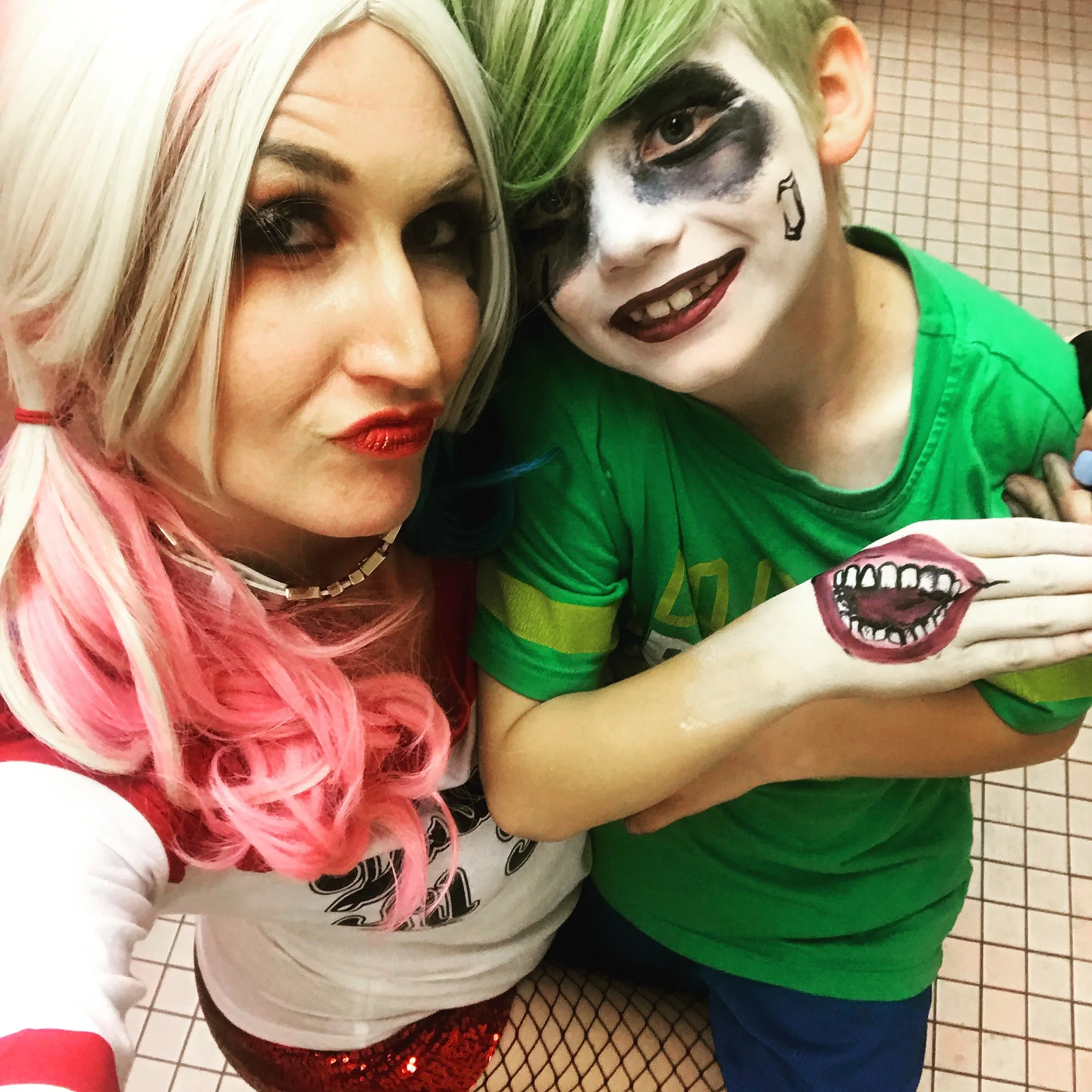 Two people dressed as Harley Quinn and the Joker in superhero costumes with face paint. They are taking a selfie in front of a tiled wall.