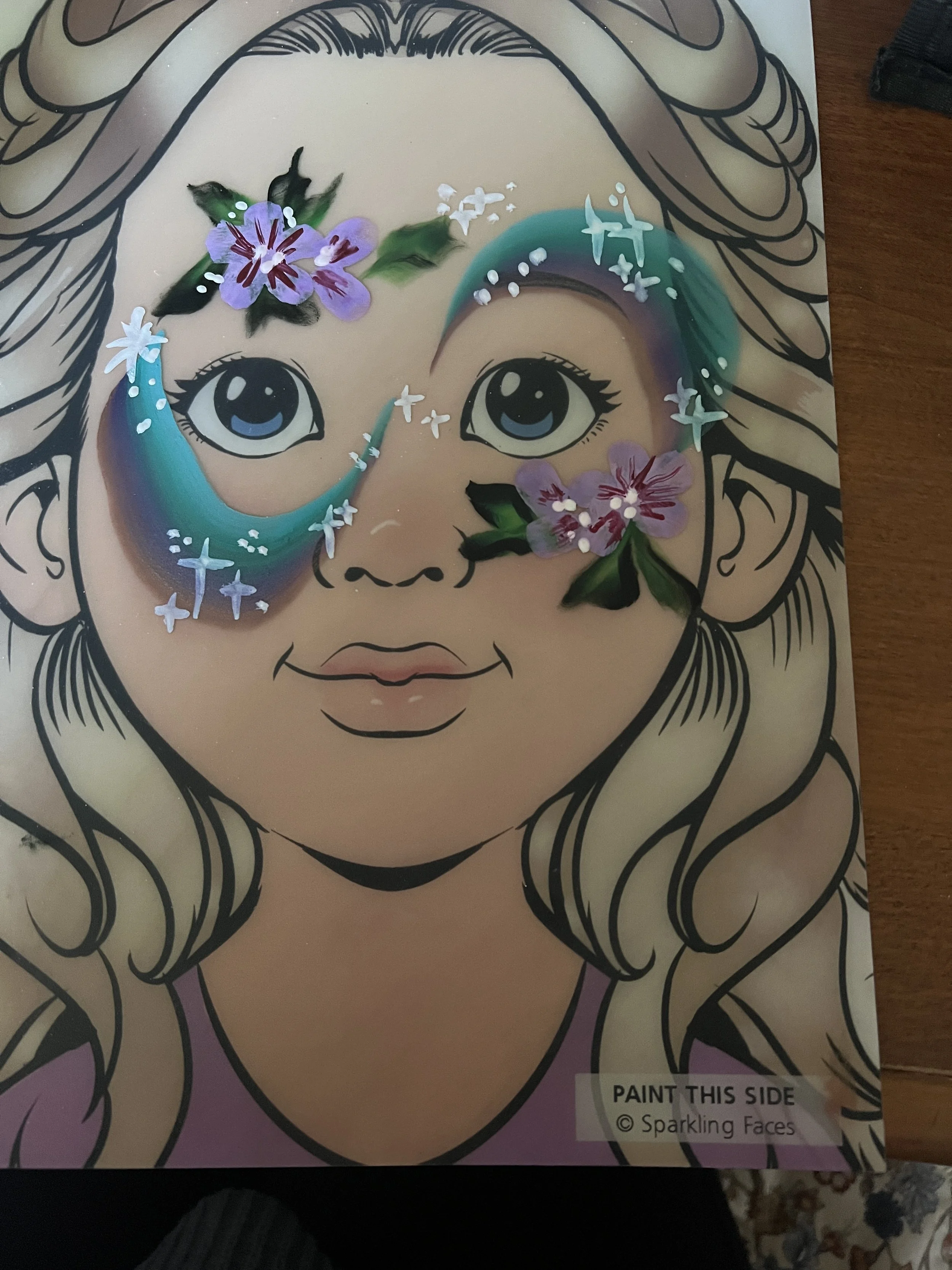 A coloring and painting activity sheet of a cartoon girl's face with blonde hair, blue eyes, a slight smile, and flowers and sparkles added around the eyes.