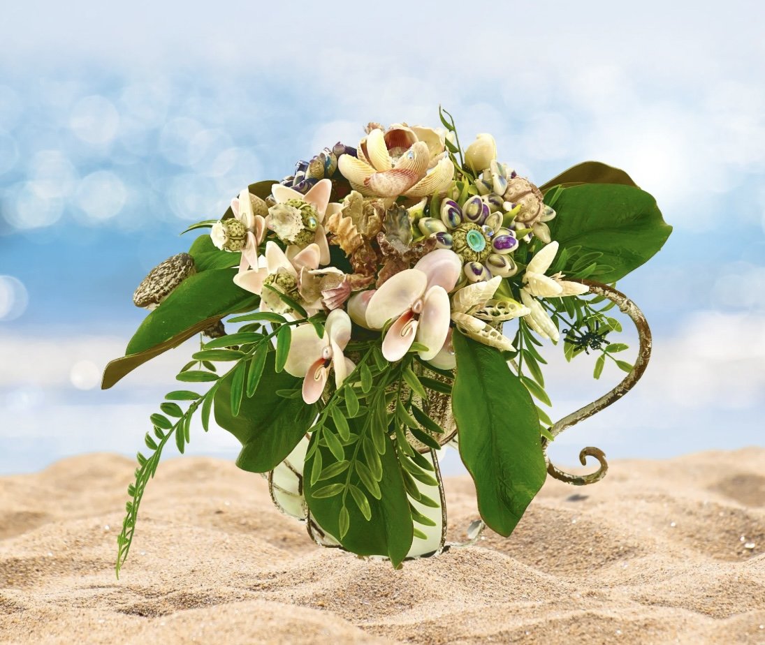 A bouquet of seashells and green foliage arranged in a clear glass vase on sandy ground with a blurred blue sky background.