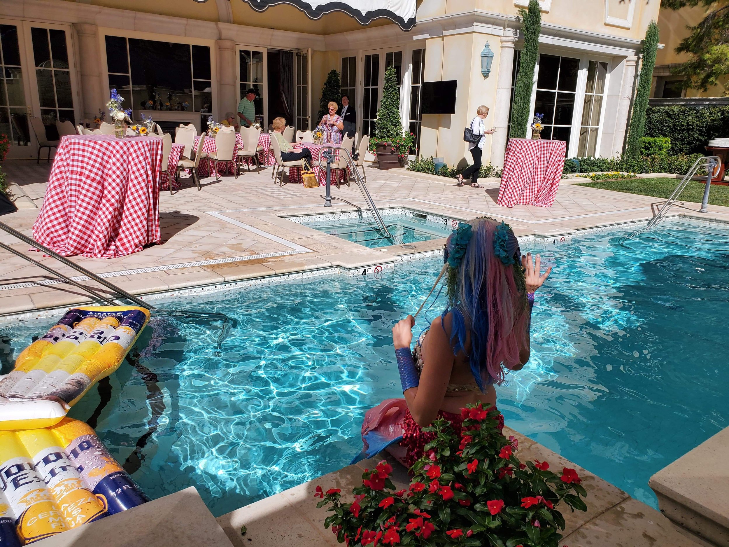 Mermaid in a pool with multi-colored hair, surrounded by red flowers, at a backyard party with guests at tables covered with red and white checkered tablecloths and a woman walking nearby.