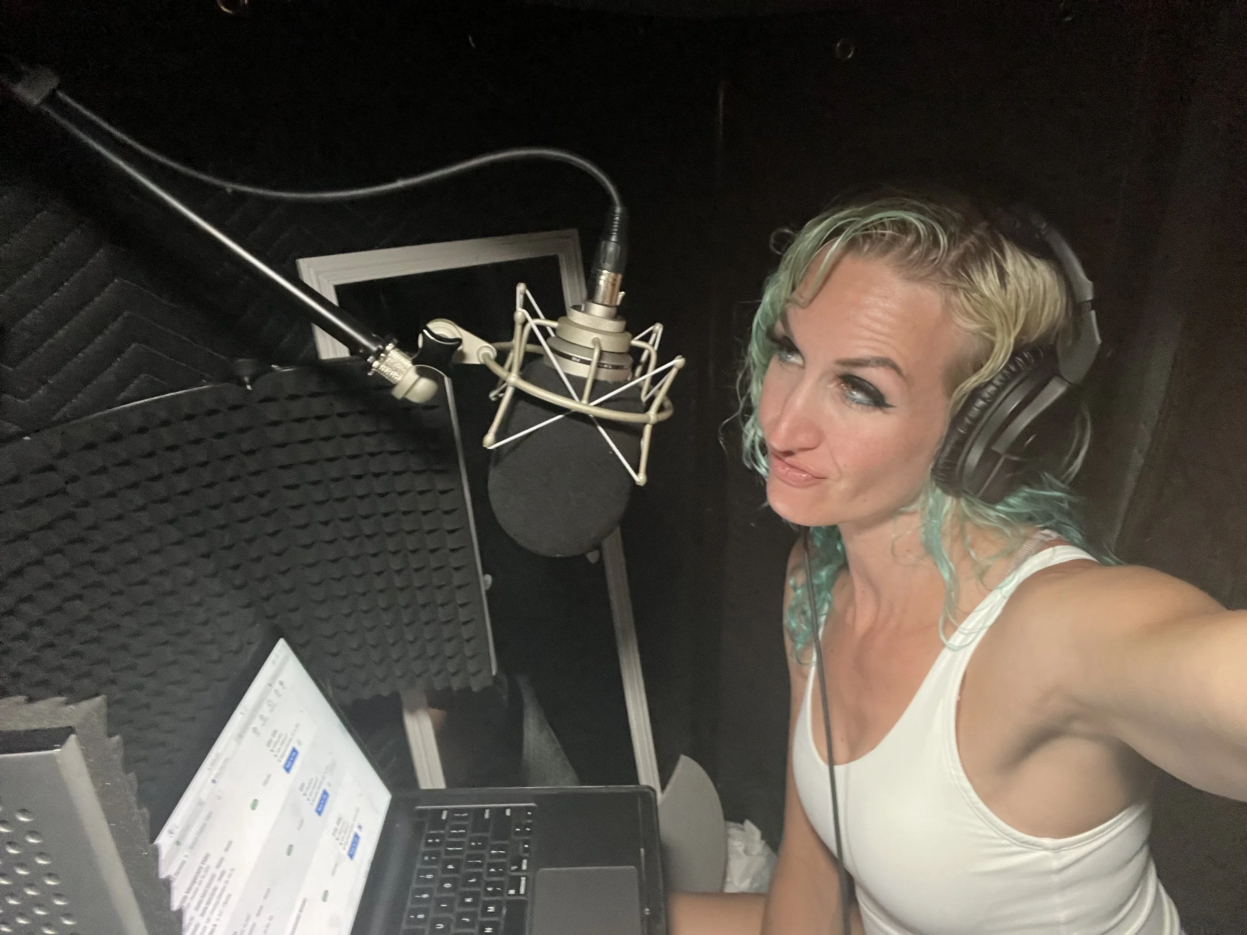 A woman with blonde hair with turquoise tips, and headphones in a recording studio, wearing a white tank top, sitting in front of a microphone, with a laptop and a monitor displaying a schedule or script in front of her. Amie Vreeken MermaidSoundCave