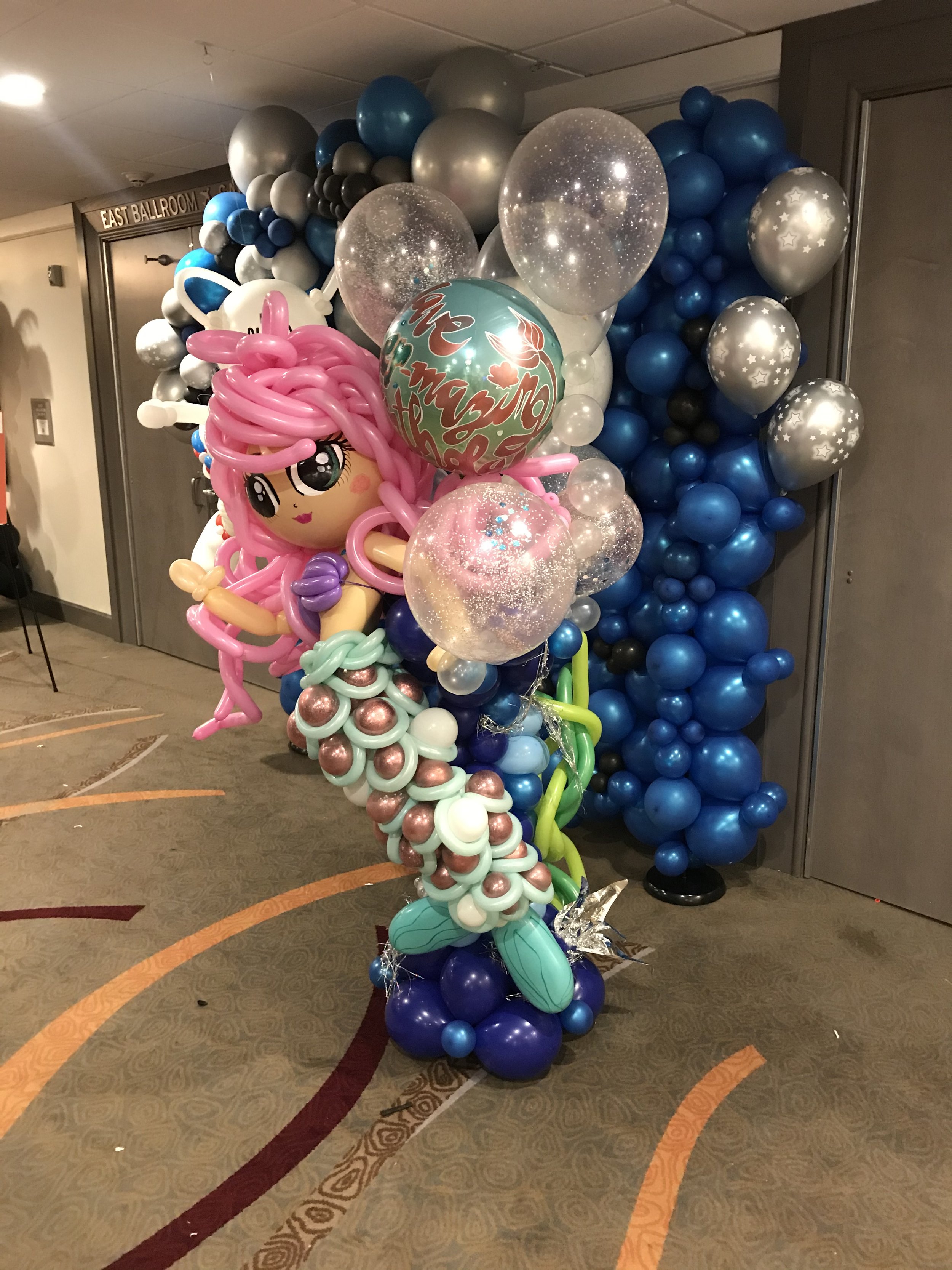 Balloon display featuring a mermaid with pink hair, surrounded by pink, purple, white, black, gray, blue, and silver balloons, with some transparent sparkly balloons, in a decorated indoor space.