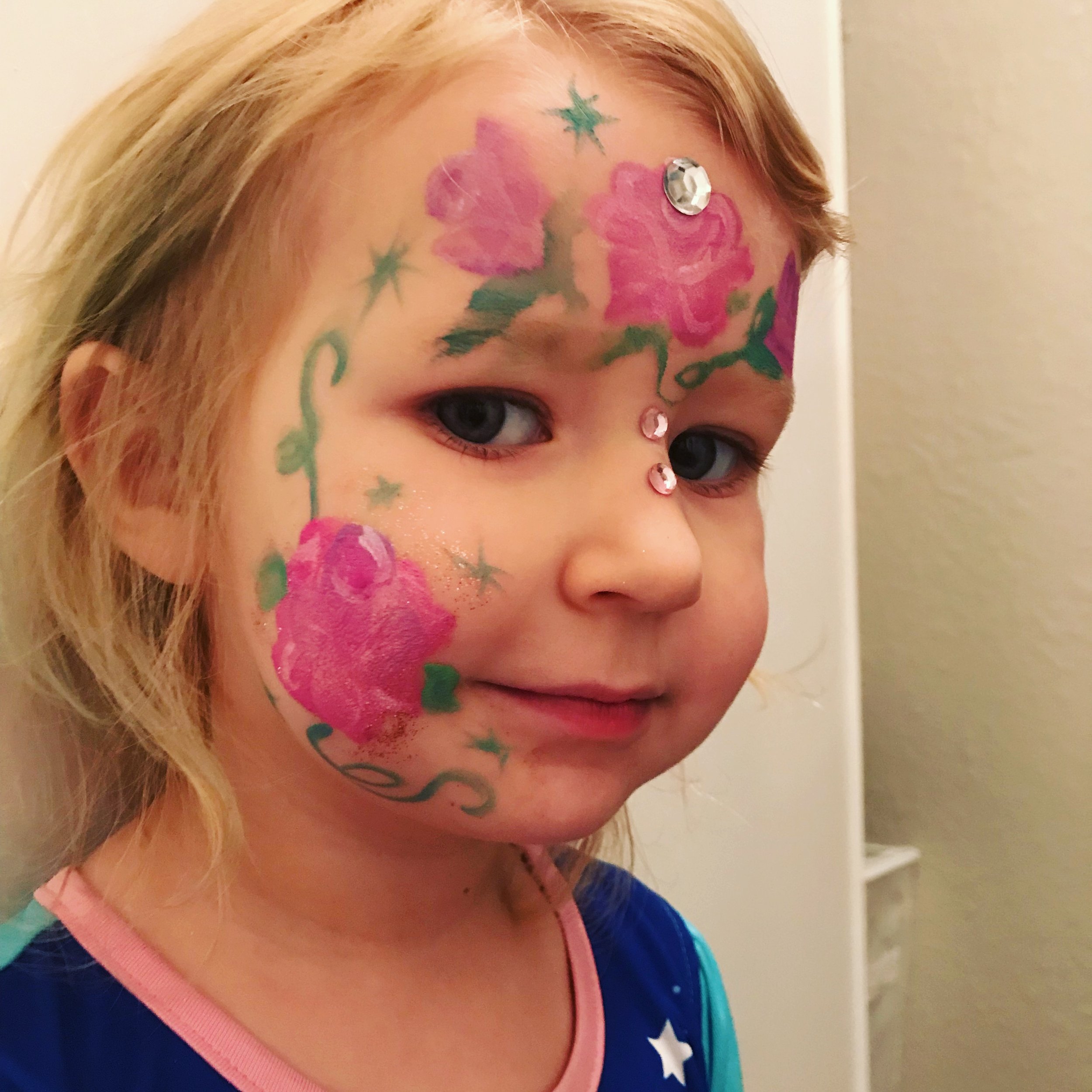 A young girl with face paint and rhinestones on her face, featuring pink flowers, green vines, and turquoise stars.