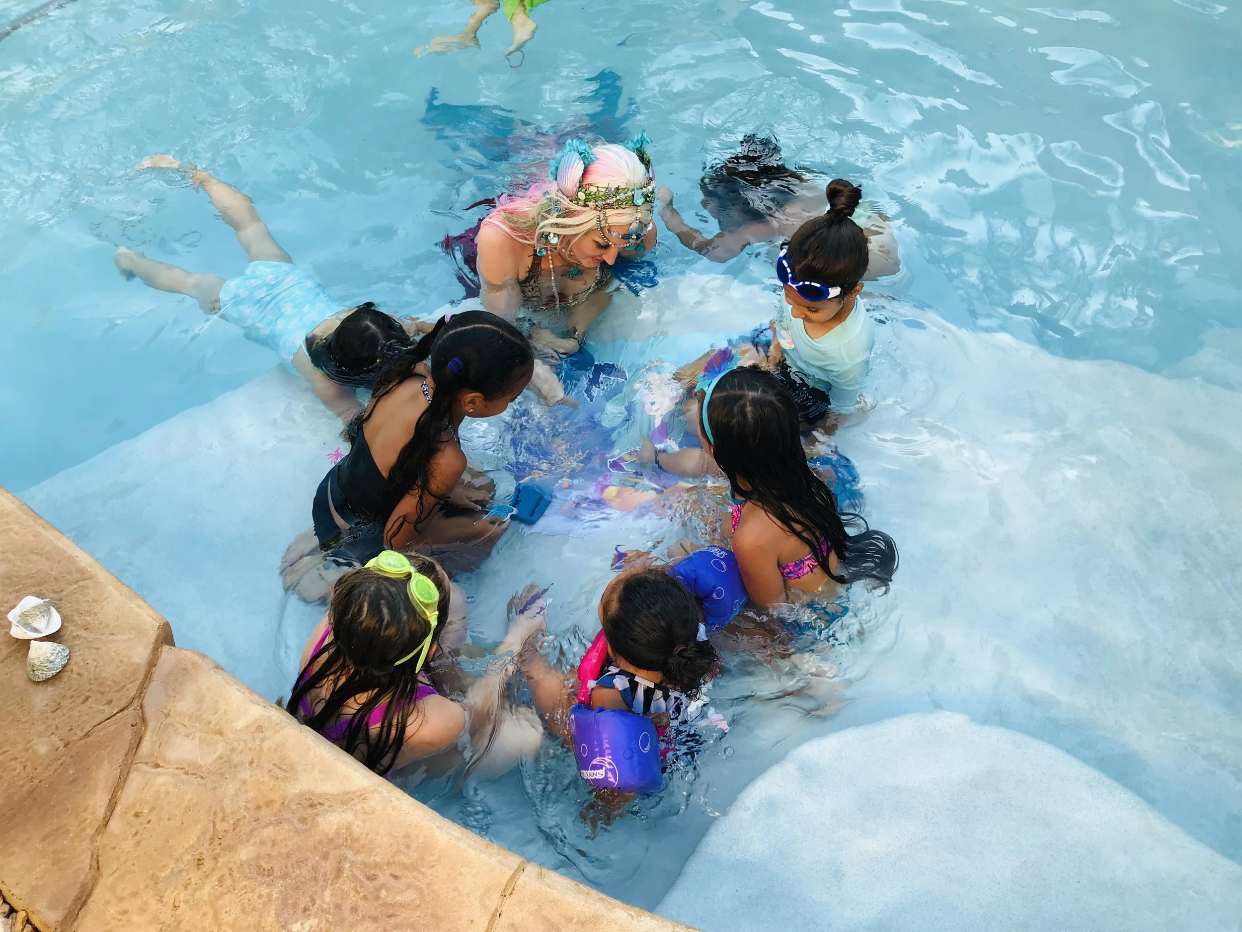 A group of children and an adult woman with colorful hair and a flower headband are in a swimming pool, playing and interacting in the water.
