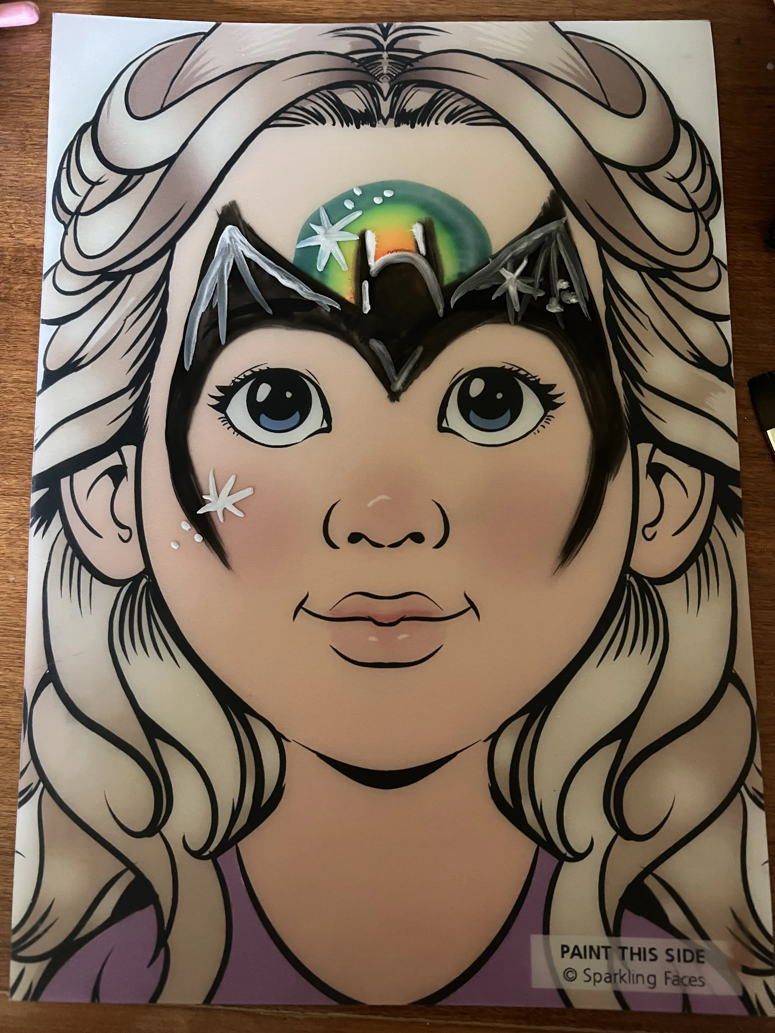 A coloring page of a girl with long wavy hair, large blue eyes, and pink lips. The top of her head has a green and orange circle with stars, and her face and hair are outlined in black with some sections filled in with color.