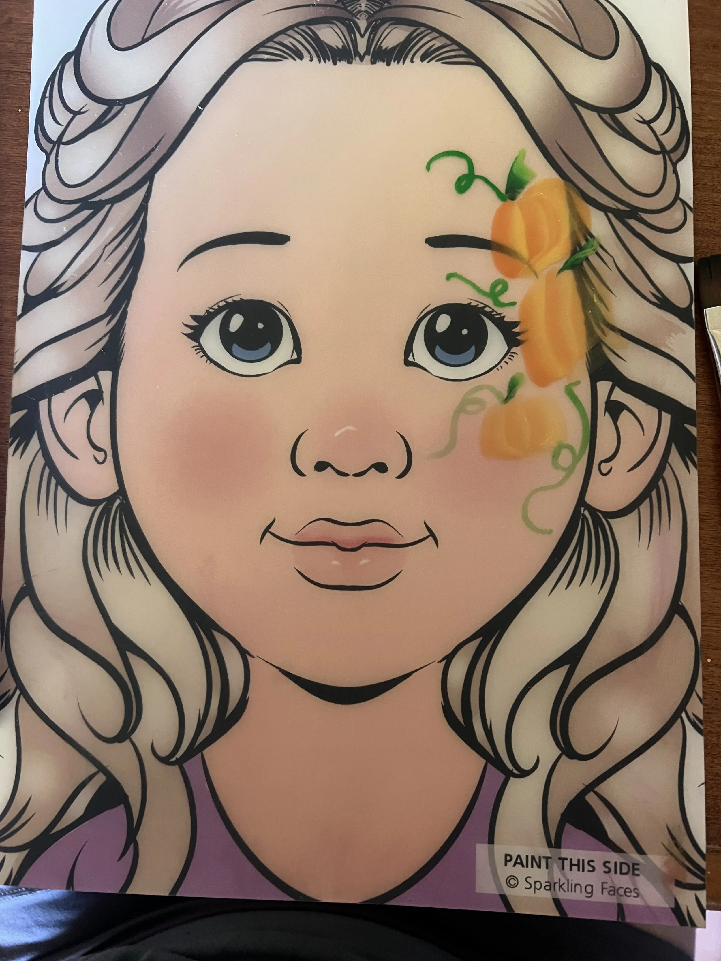 A coloring page of a young girl with large blue eyes, curly hair, and a cheerful expression. The page has some orange and green paint splashes on the right side, resembling a pumpkin and vines.