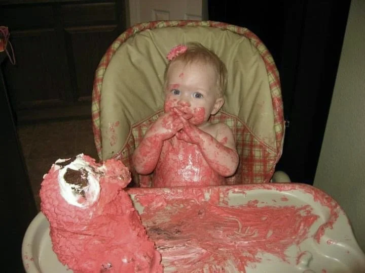 One year old girl on her birthday covered in bright red frosting with a half eaten 3D Elmo from Sesame Street cake sitting on the high chair tray.