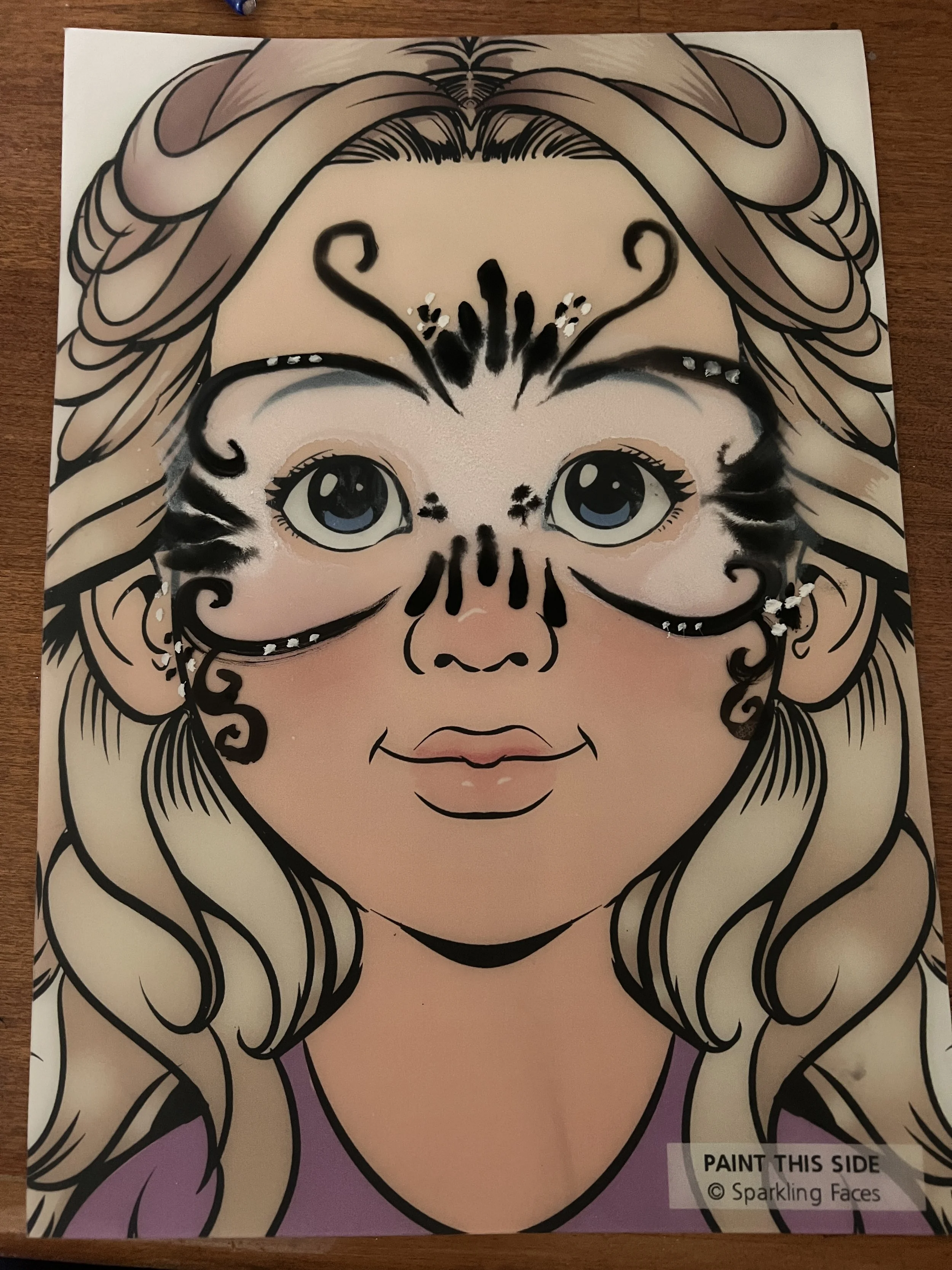 A coloring sheet of a girl with long, wavy blonde hair and blue eyes, decorated with a black face mask design, requiring coloring in. The sheet says 'Paint This Side' at the bottom right corner.