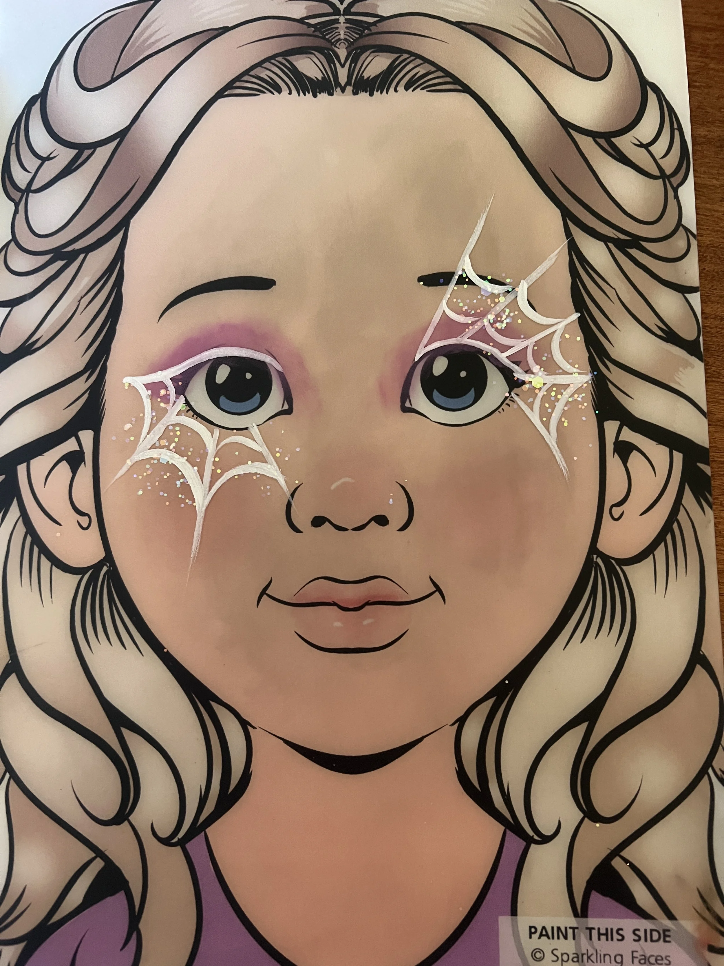 A cartoon drawing of a girl with blonde curly hair, big blue eyes, and pink makeup. There are white spiderweb designs with glitter near her eyes, suggesting a Halloween theme.