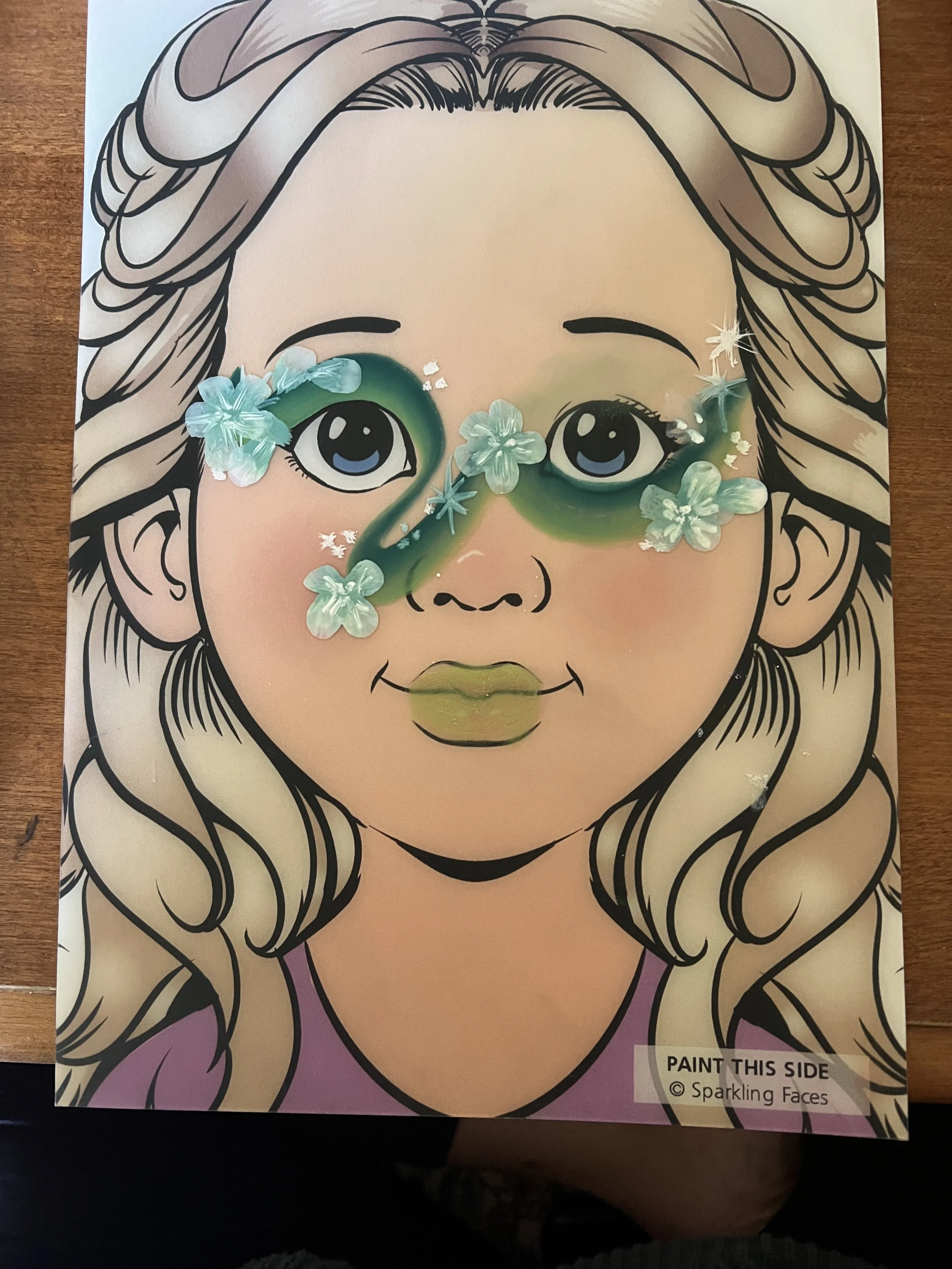 A coloring page of a girl with long, wavy hair and large eyes, with green and pink lips. The page is being decorated with small blue flowers and star-shaped sequins placed across the face to create a sparkling effect.