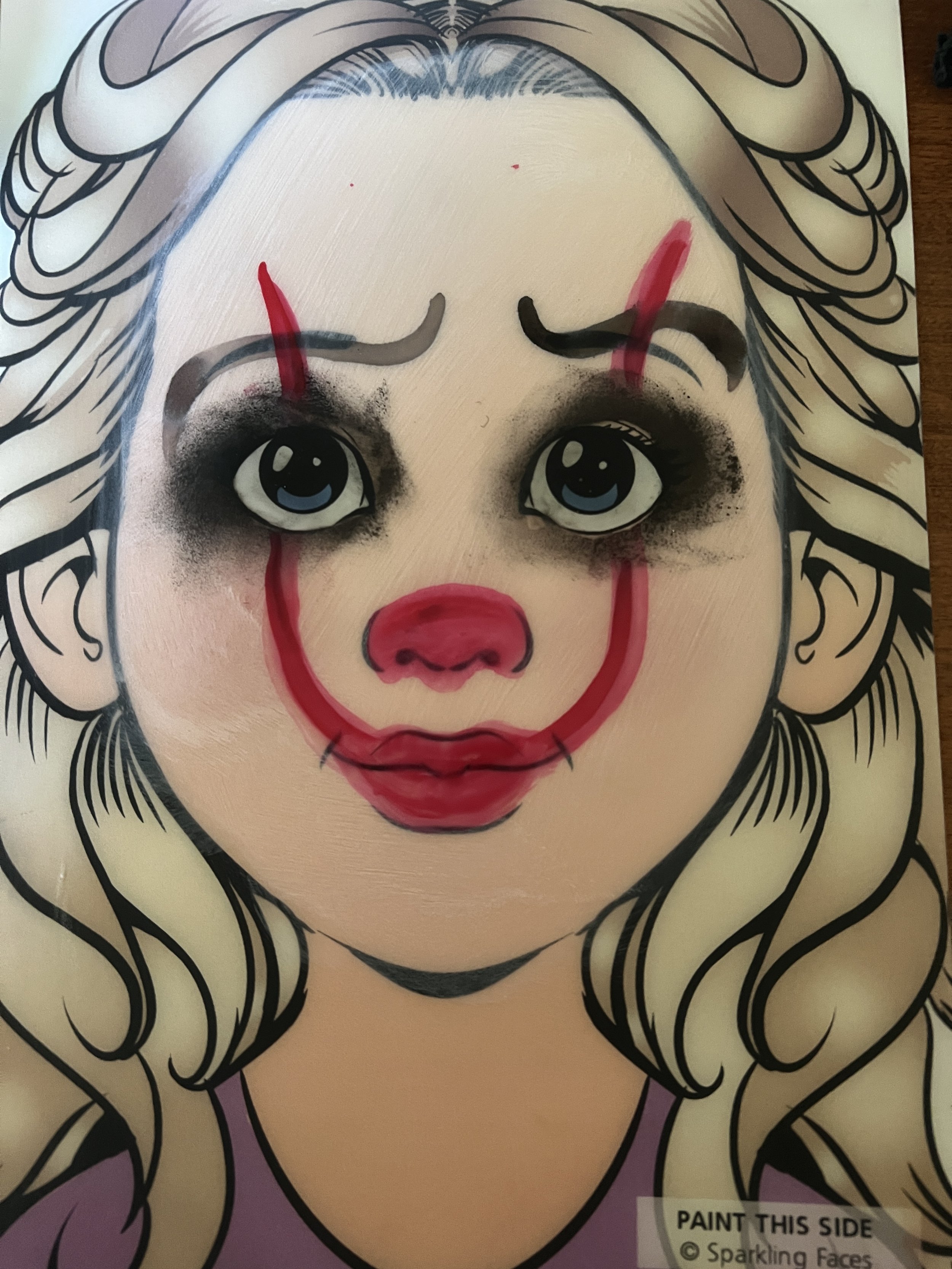 A coloring book illustration of a girl with long blonde hair, overlaid with red, black, and blue clown makeup, including a red nose, red lips, black eyeshadow, and red lines above her eyebrows.