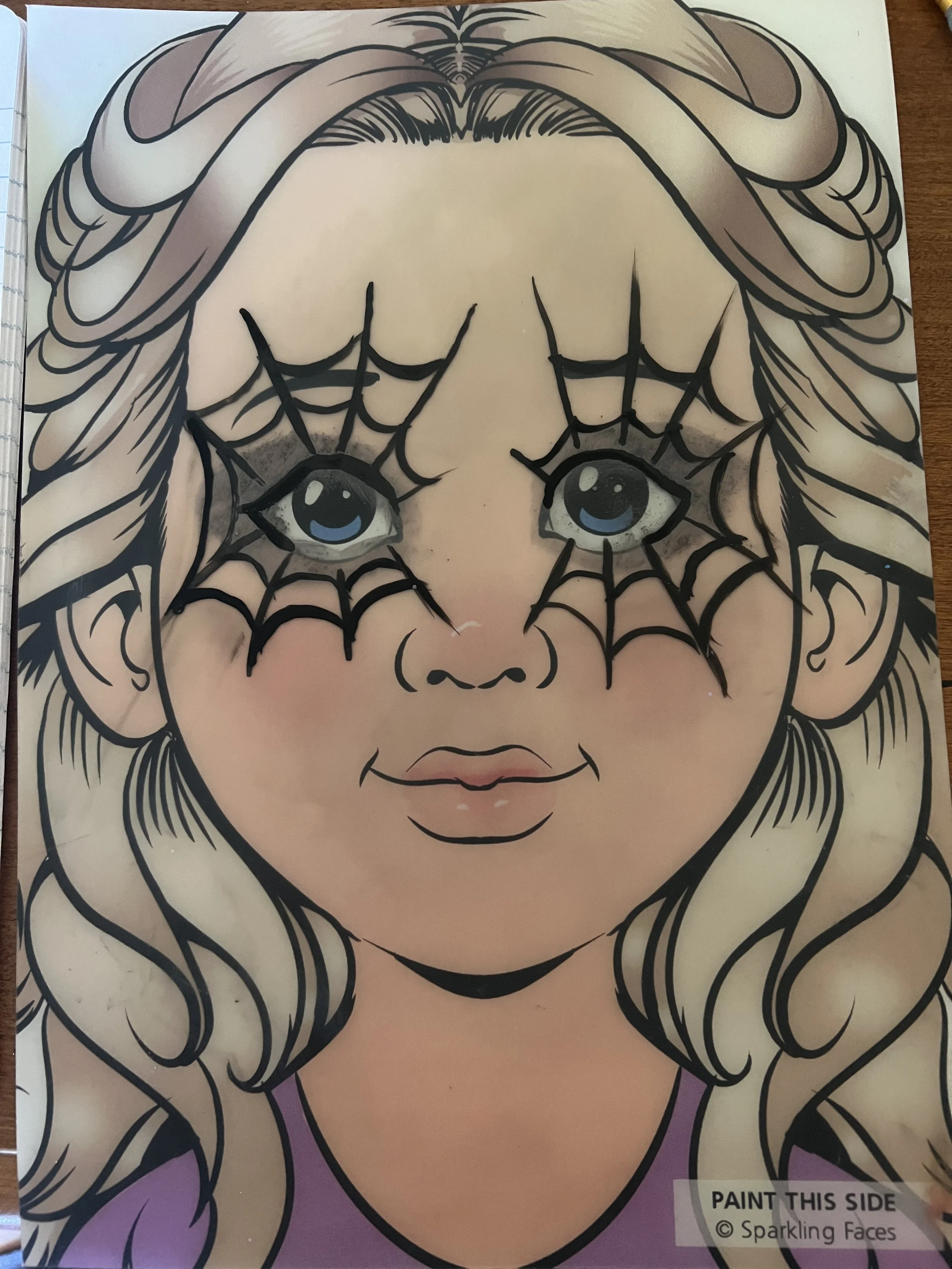 Child with blonde hair and blue eyes, face painted with black spider web design over the eyes.