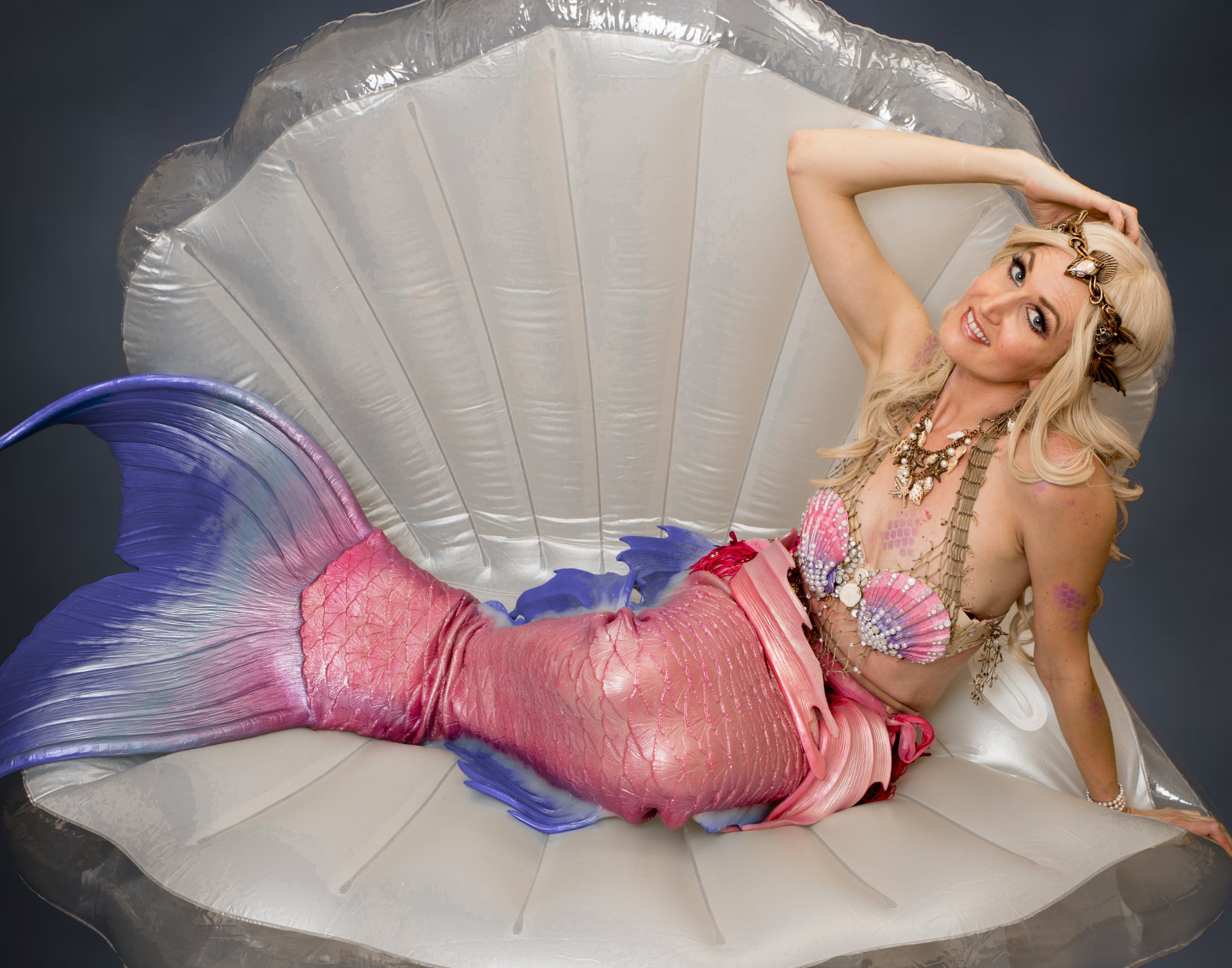 A woman dressed as a mermaid, with a pink and purple tail and seashell top, lounges on a large faux clam shell with a dark background.