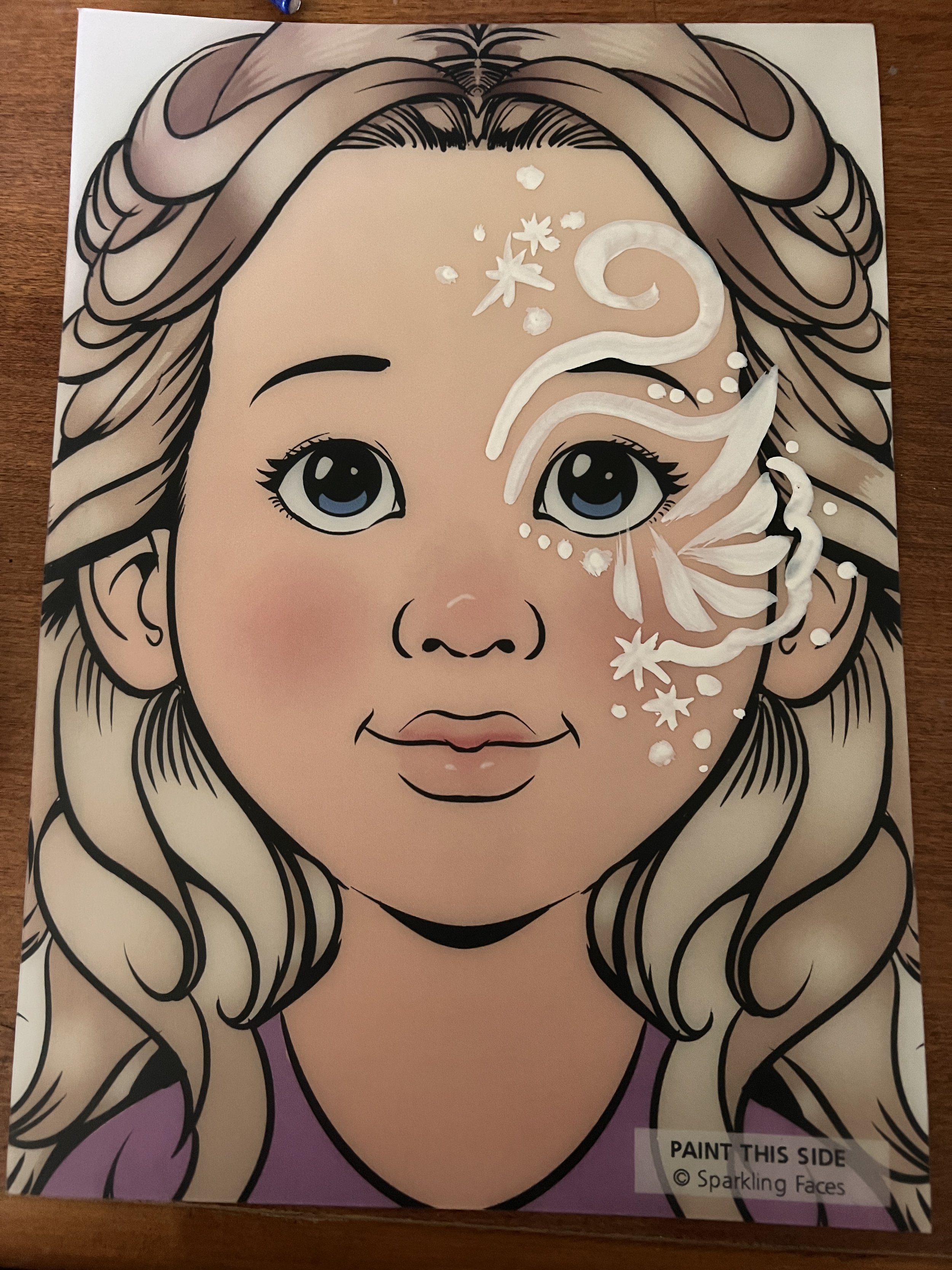 Coloring sheet of a girl with long wavy hair, large blue eyes, and pink cheeks, with white paint or marker designs on her face, including swirls and small star-like shapes, and a label that says 'Paint This Side © Sparkling Faces'.