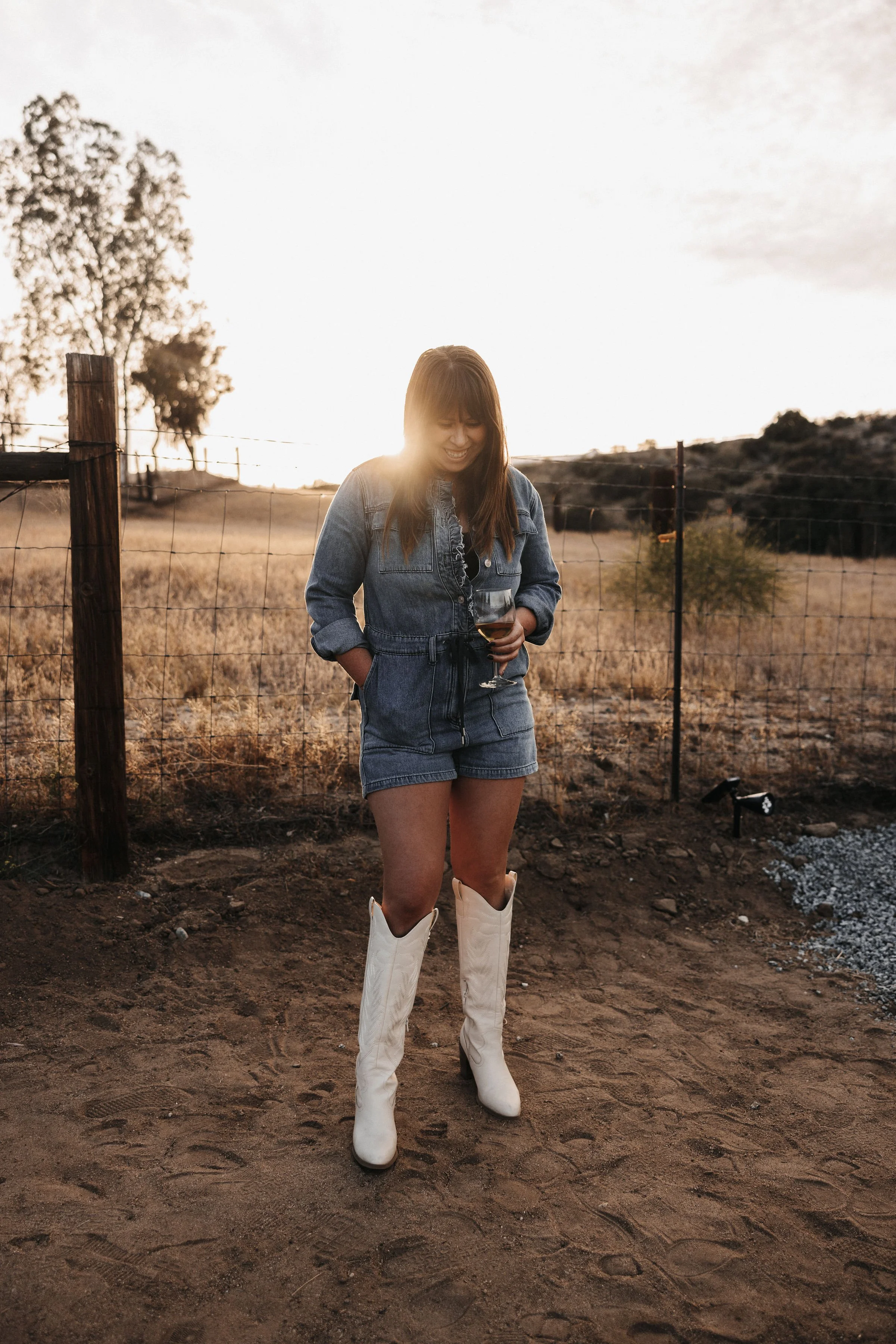 Young woman in denim jacket and shorts with white cowboy boots holding a wine glass, standing outdoors on dirt ground during sunset.