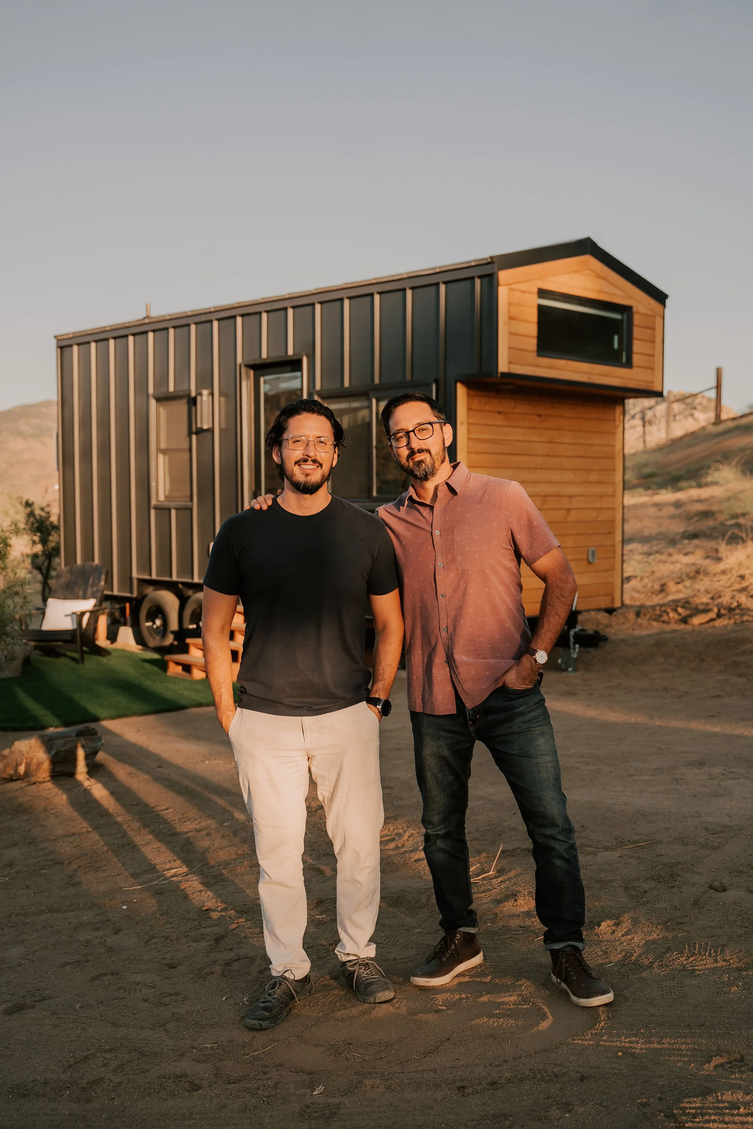 Two men standing outdoors in front of a modern tiny house with a metal and wood exterior, smiling at the camera during sunset.