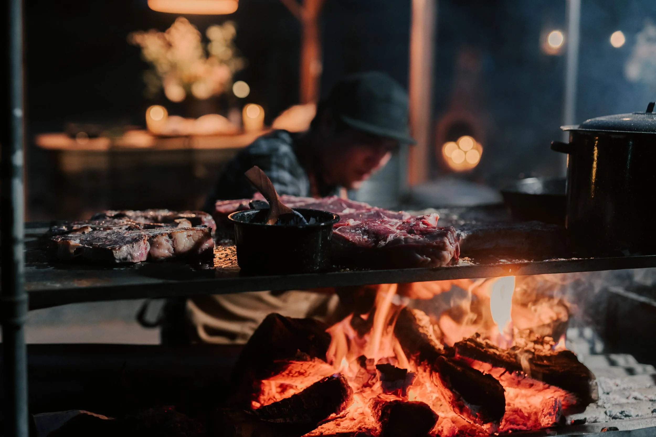 Outdoor grilling scene with steaks cooking over a wood fire.