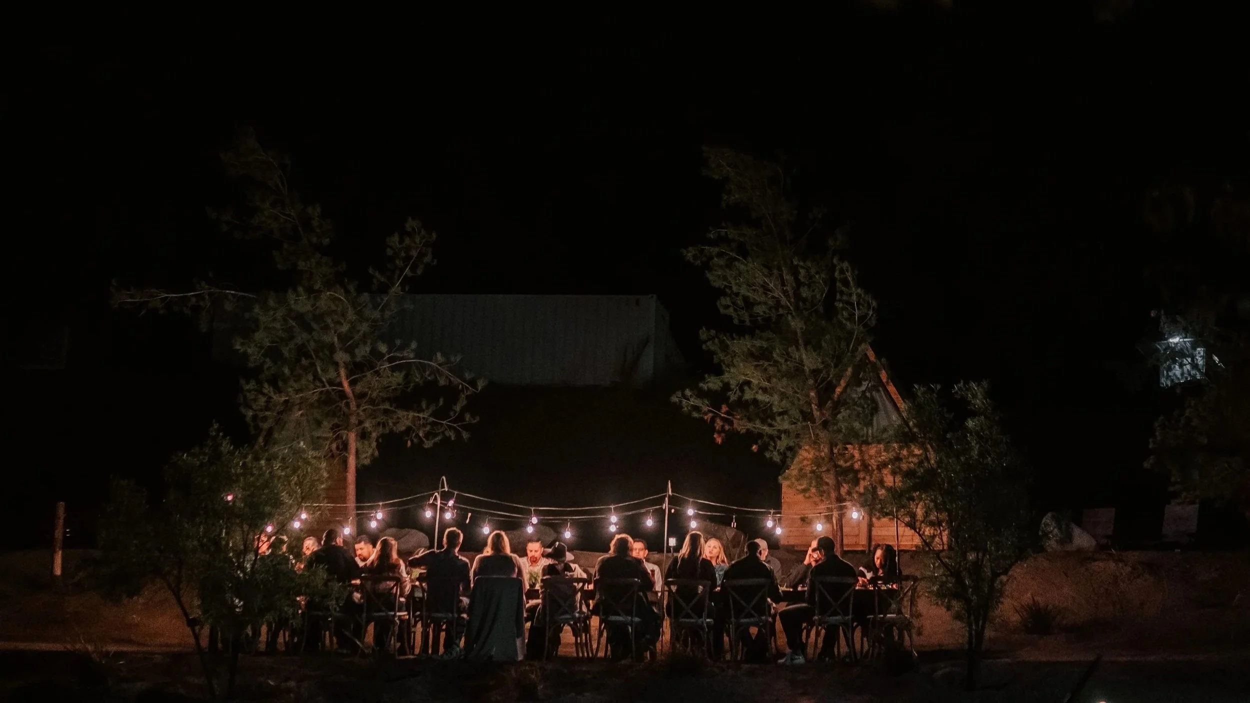 Nighttime outdoor gathering with a group of people sitting at a long table under string lights, surrounded by trees and rustic buildings.