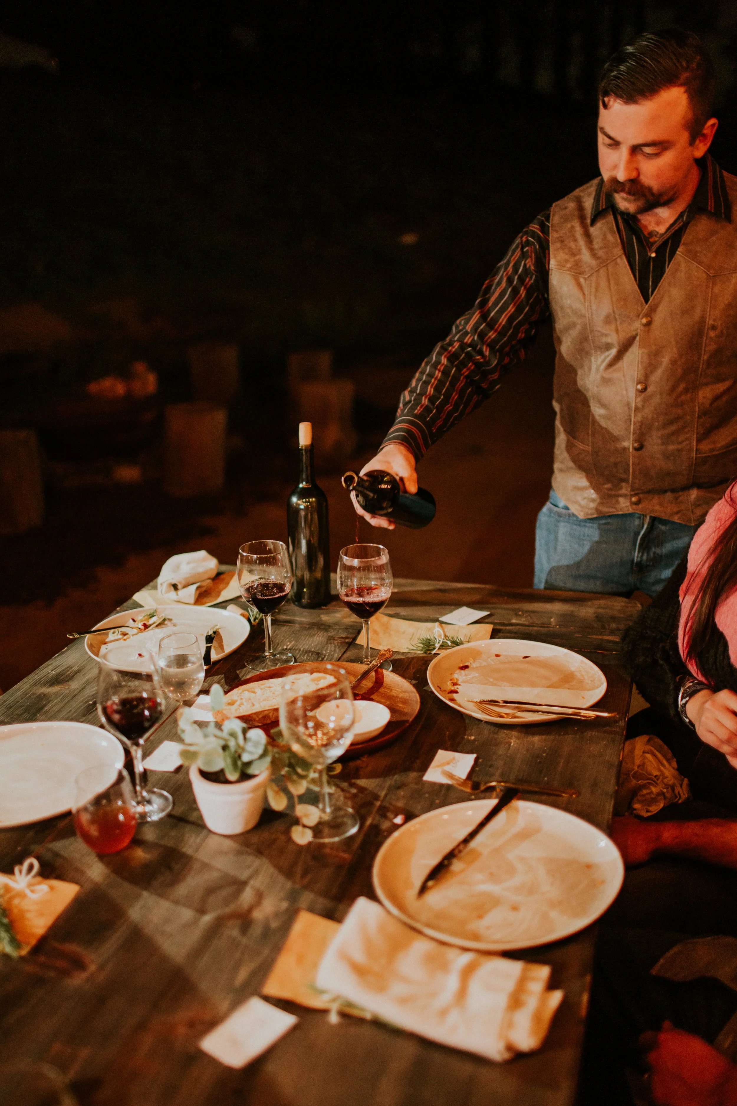 A man pours red wine at a dinner table set outdoors at night, with empty plates, wine glasses, and a potted plant on the table.