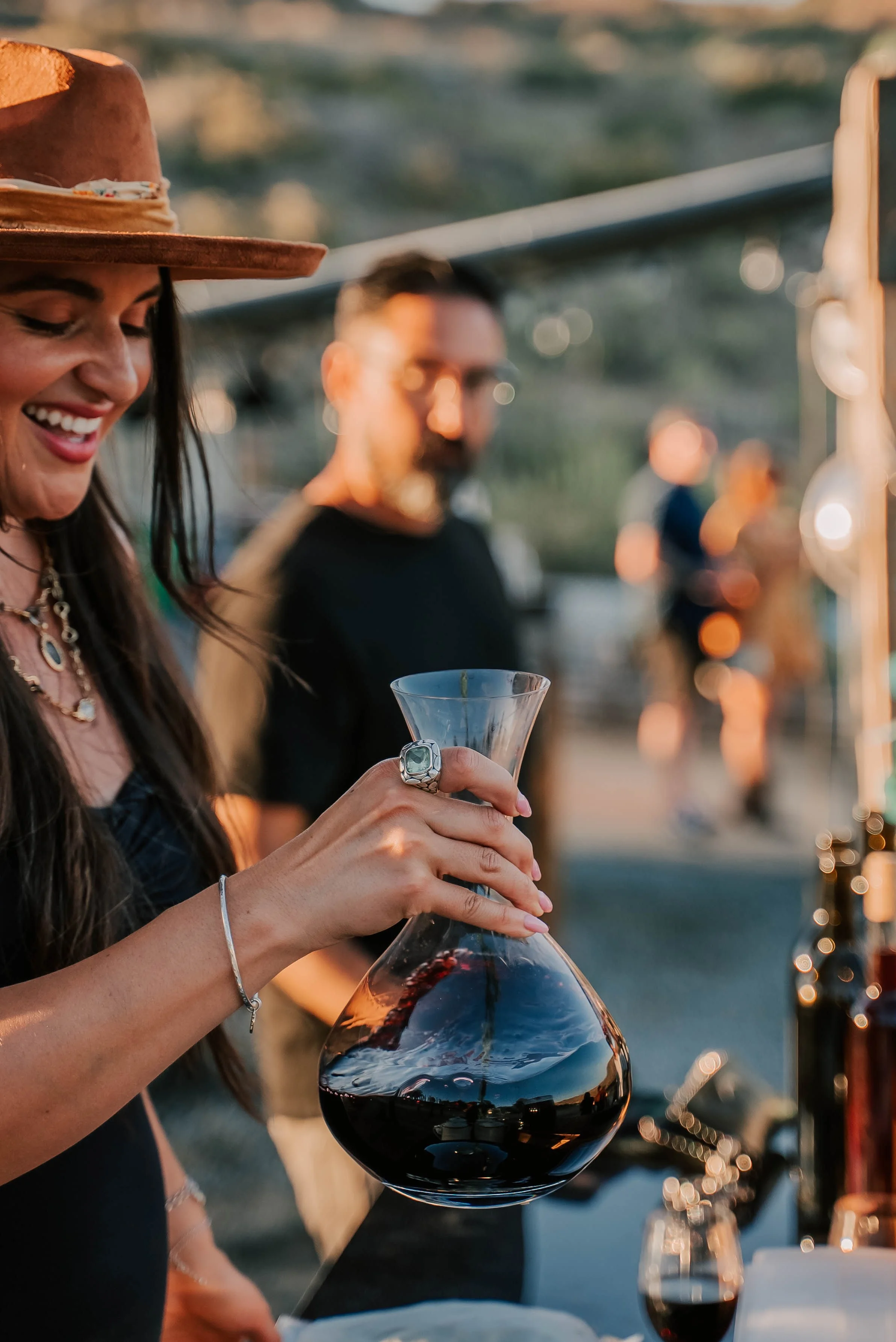 Woman smiling and holding a glass pitcher of red wine at an outdoor event during sunset, with other people blurred in the background.