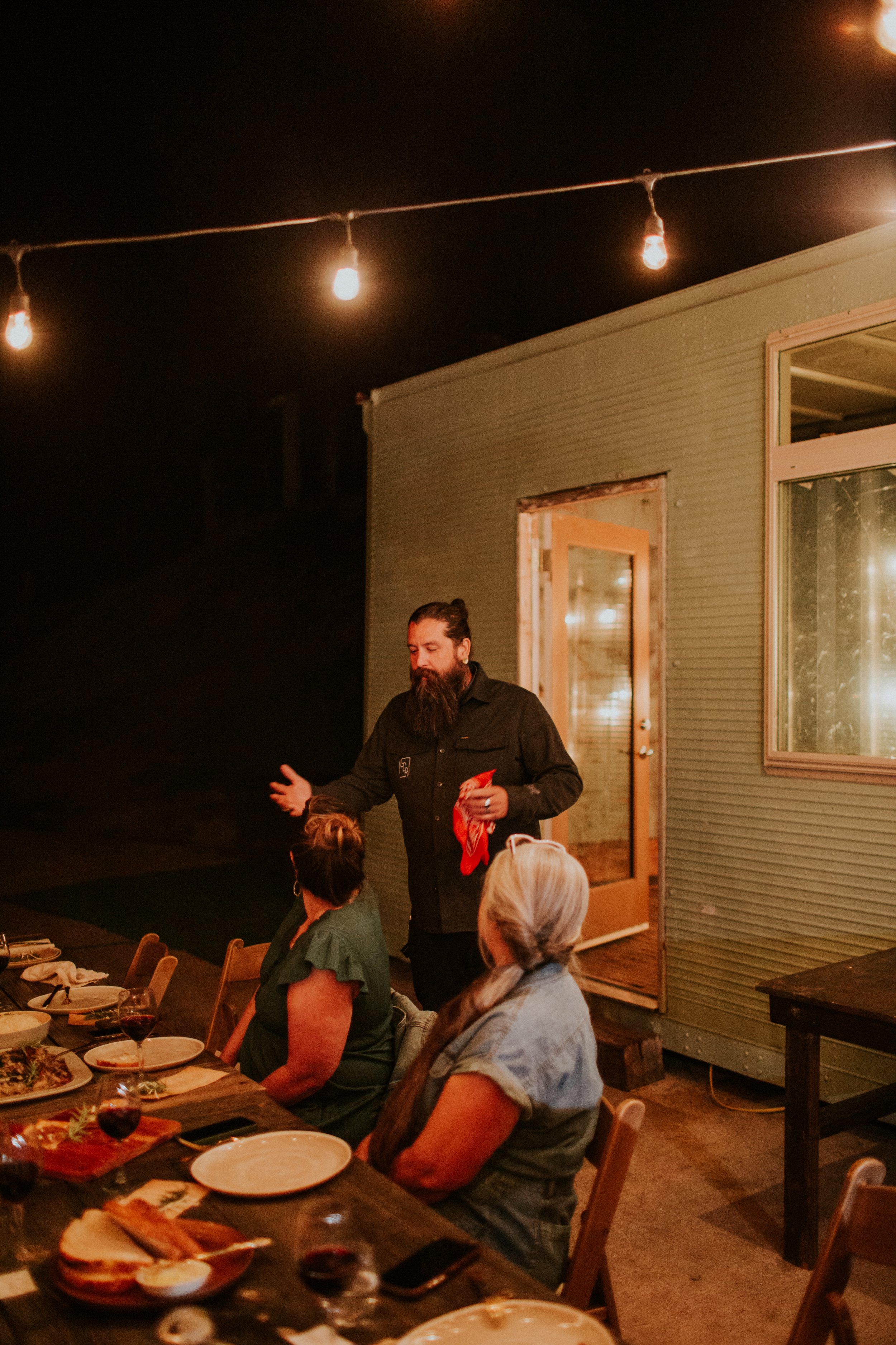A man with a beard, wearing a black shirt, is speaking to two women sitting at a dinner table outdoors under string lights at night. The table has plates of food and glasses of red wine.