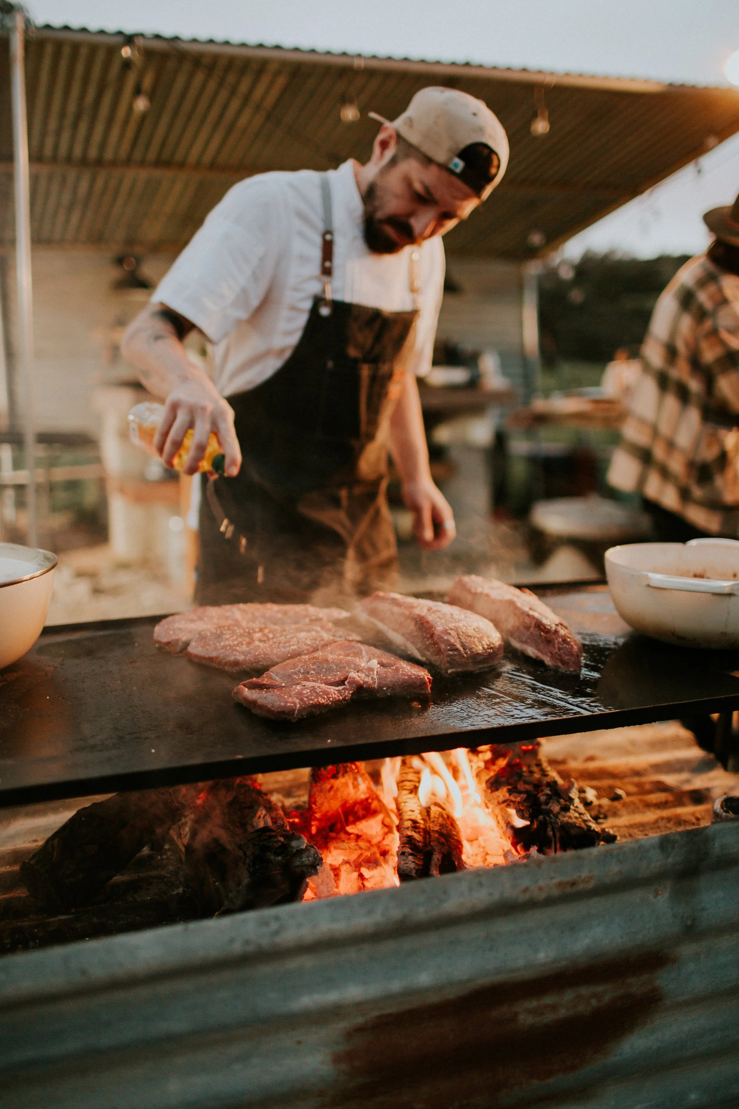 Man grilling several steaks on an outdoor grill at sunset.