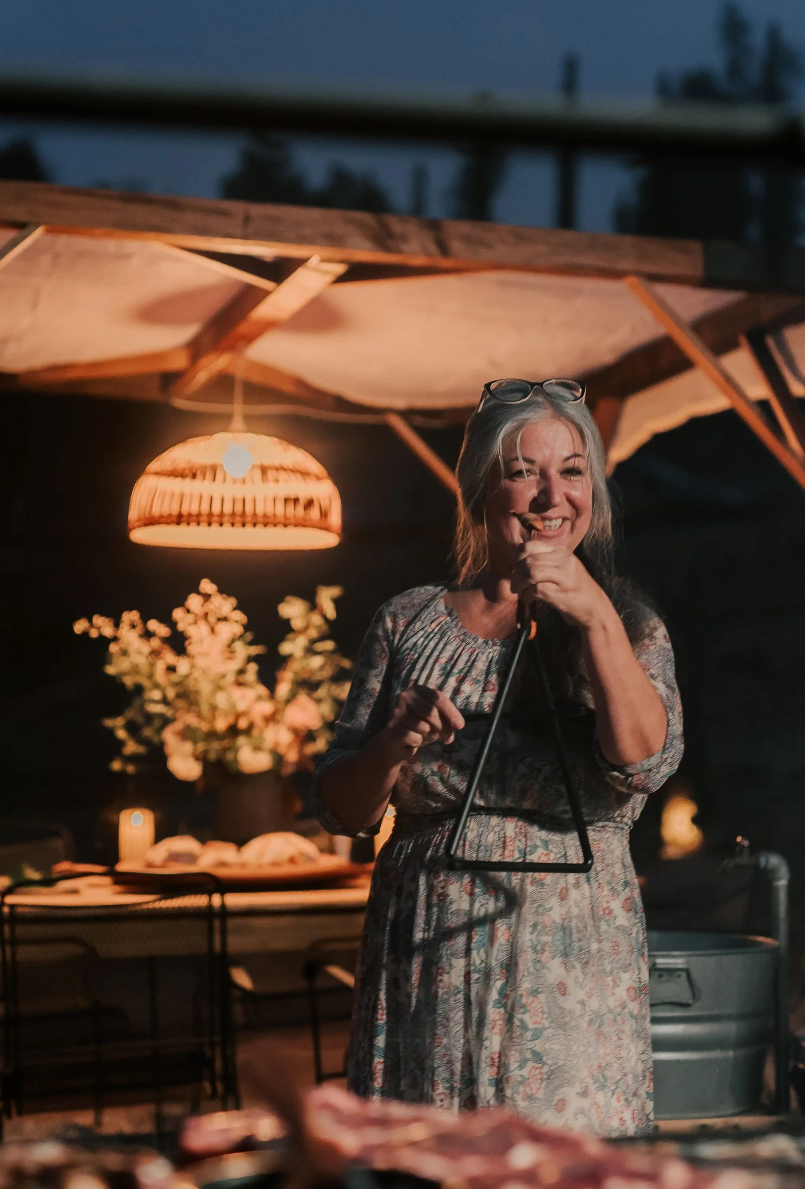 A woman with gray hair and glasses on her head smiling and holding a microphone at an outdoor evening event, with warm lighting and a flower arrangement in the background.