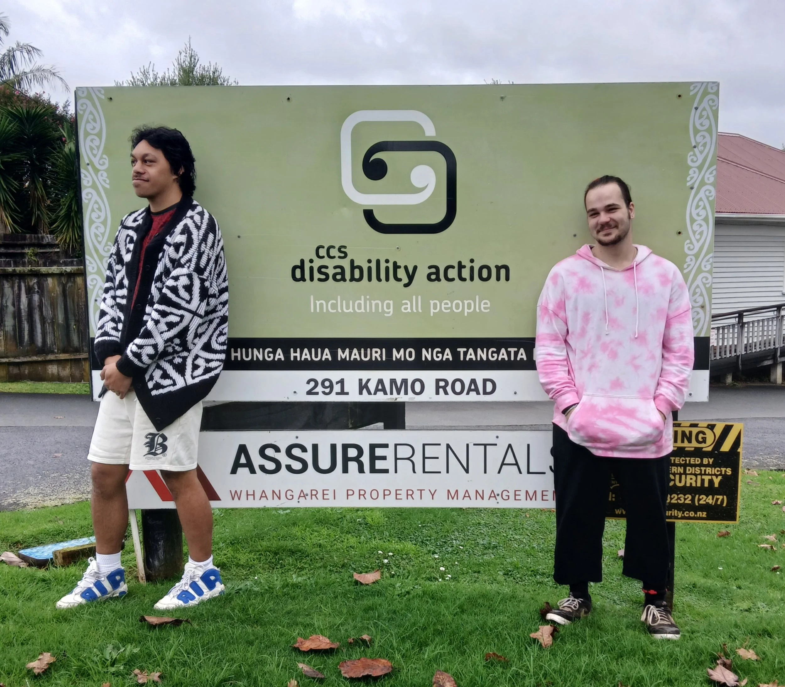 Shannon and Rus, two young men, stand in front of the CCS Disability Action sign.