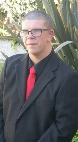 Portrait photo of Matthew wearing a black suit with red tie and glasses.