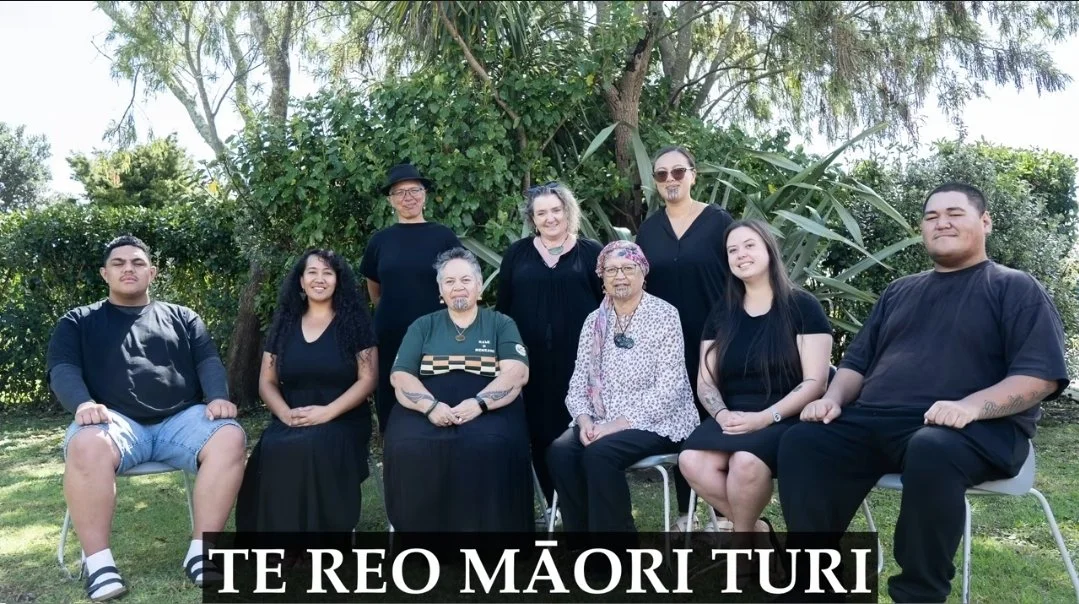 A group of people look to the camera. They are in two lines, with one group of six people seated in the front and three people standing at the back. They are outside and all smiling at the camera.
