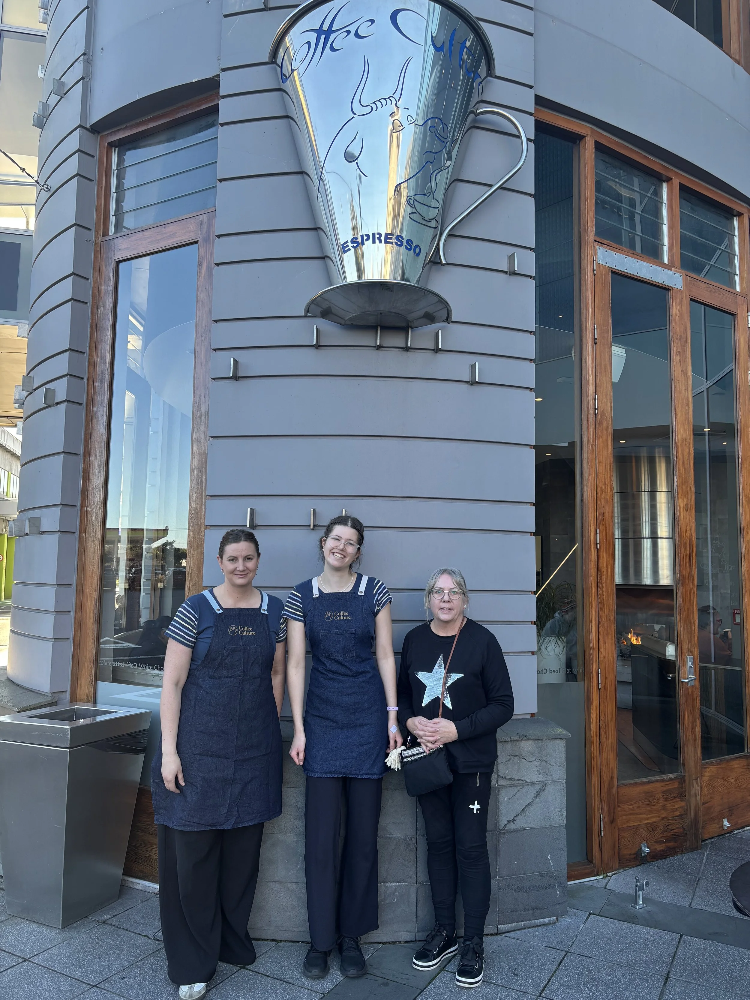 Three women stand outside a cafe building, smiling.