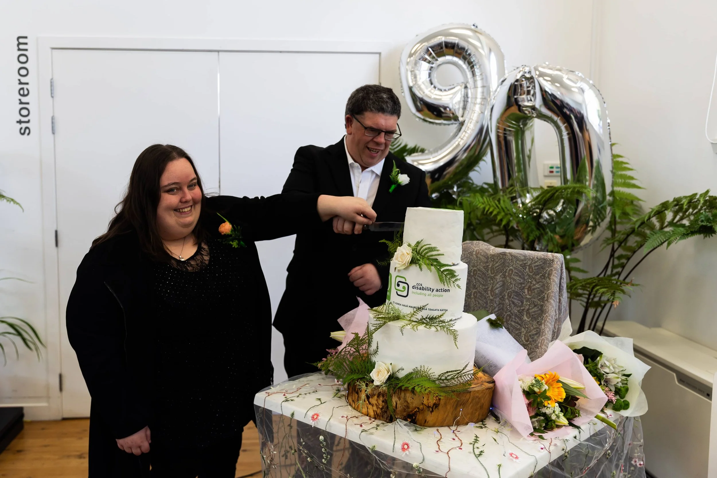 Amanda cutting into a three tier cake decorated with the CCS Disability Action logo. Behind her are silver balloons in the shape of 9 and 0.