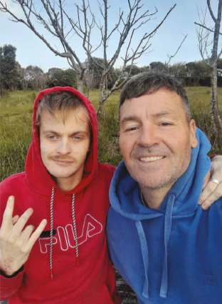 Selfie of Blake and Tim outdoors, smiling. Blake is wearing a red hoodie and making a sign with one hand. Tim is in a blue hoodie.