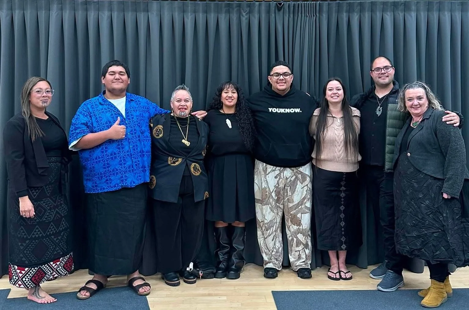 A group of seven people stand together. They are smiling and have their arms around each other, except one person who is holding his hands in a thumbs up gesture.