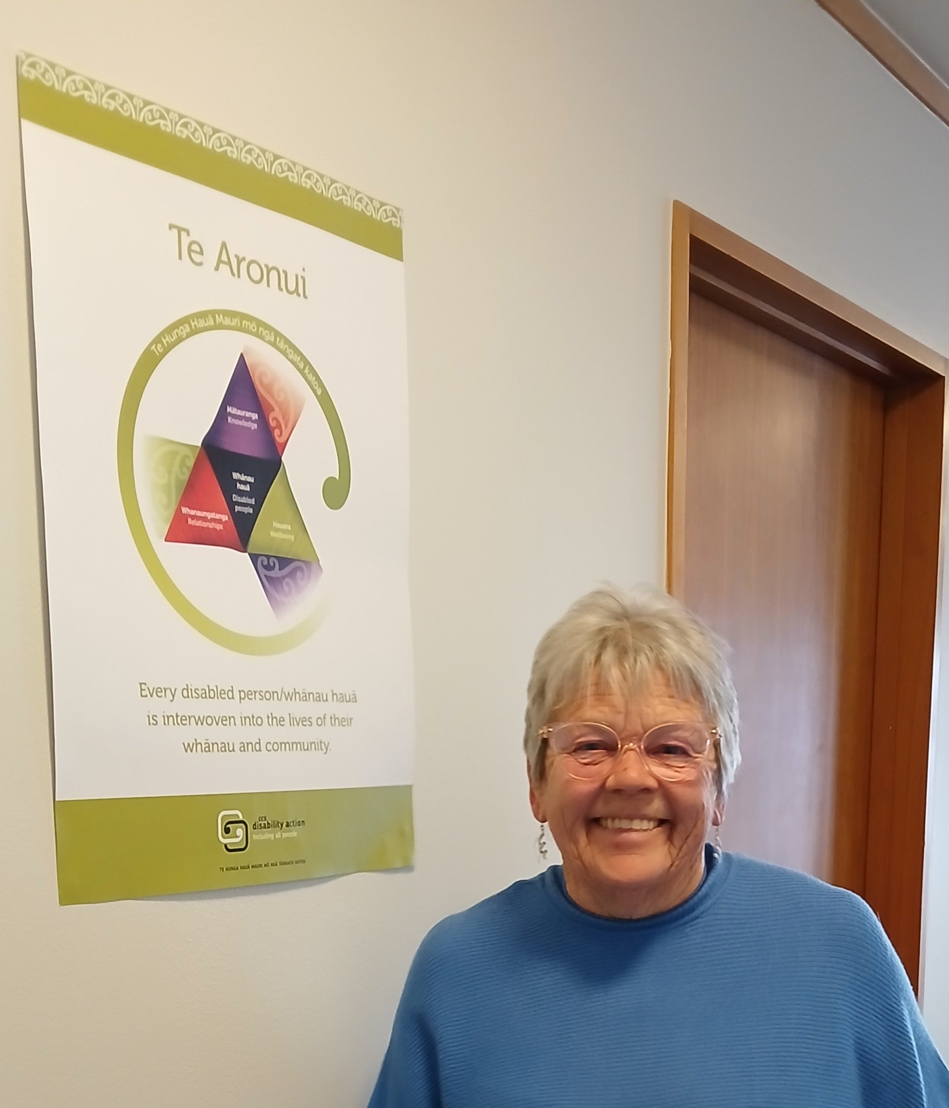 Woman with short grey hair and glasses beside a poster of Te Aronui.