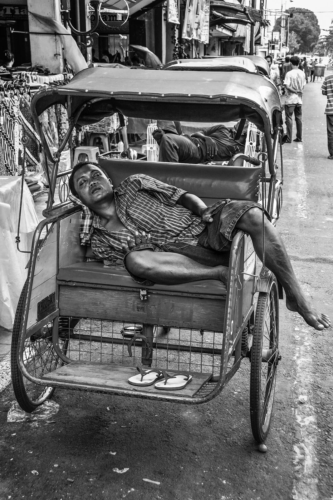 Photographie de rue noir et blanc : un jeune homme dort confortablement installé à sur le siège de son rickshaw. Style contrasté signé Hugues Hardy.