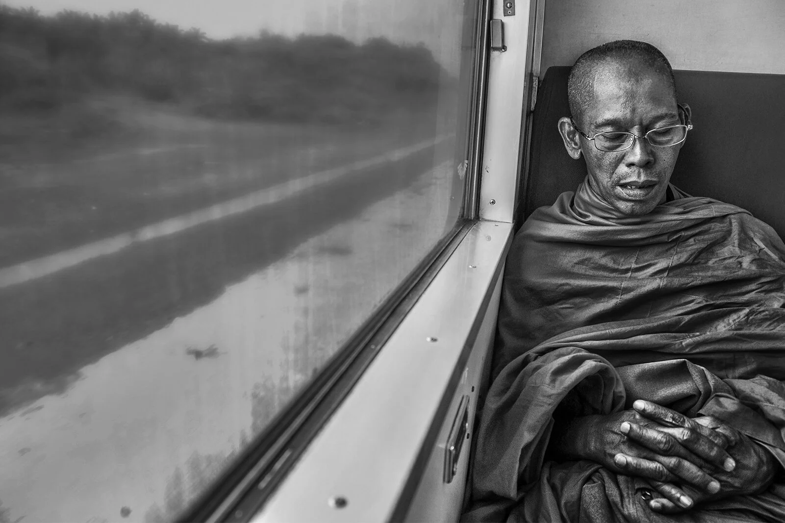 Portrait noir et blanc d'un moine bouddhiste thaïlandais assoupi dans un train en mouvement. Composition sobre et contrastée.