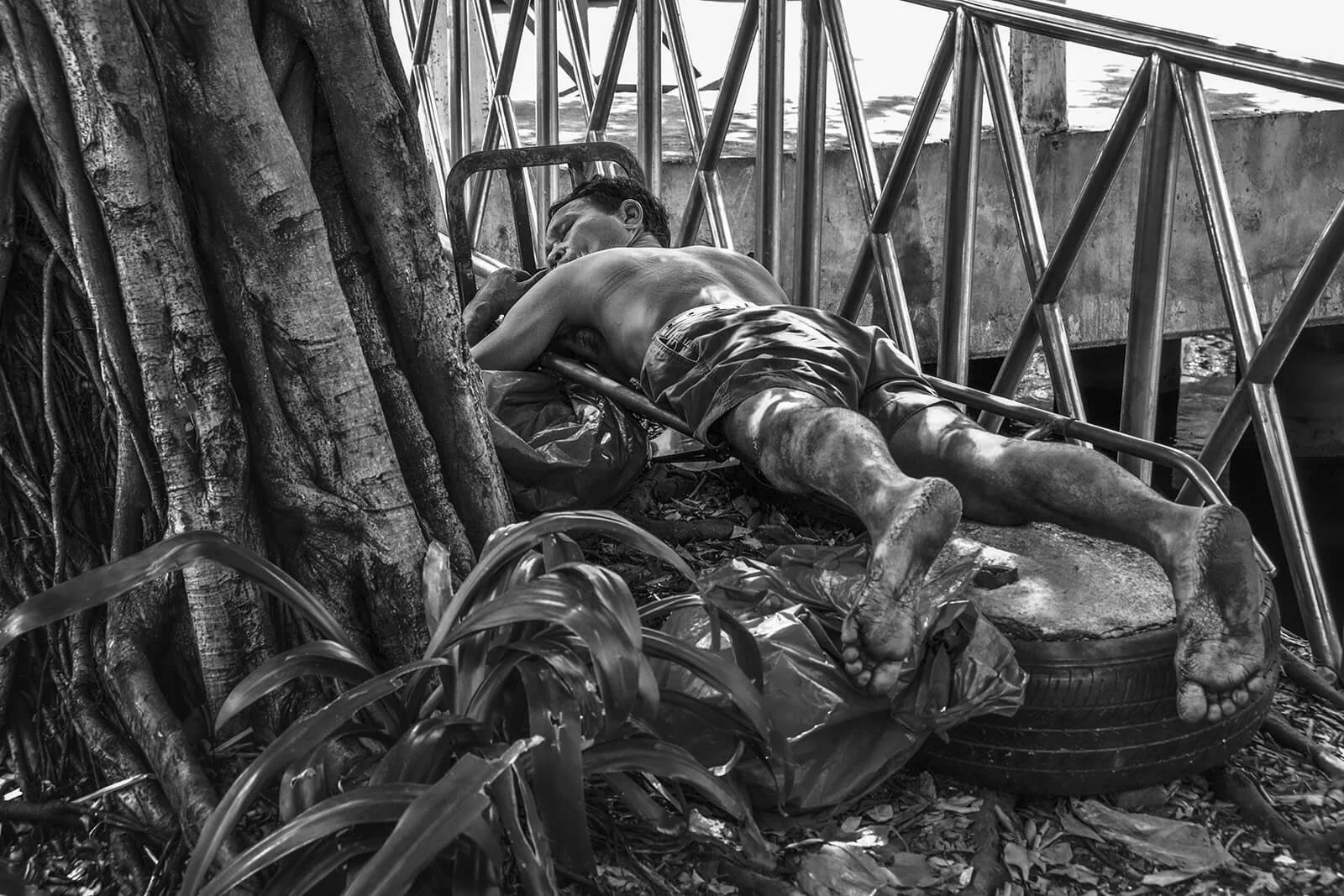Image de rue insolite : un homme dort à plat ventre sur un diable de transport sous un arbre. Noir et blanc, Hugues Hardy.