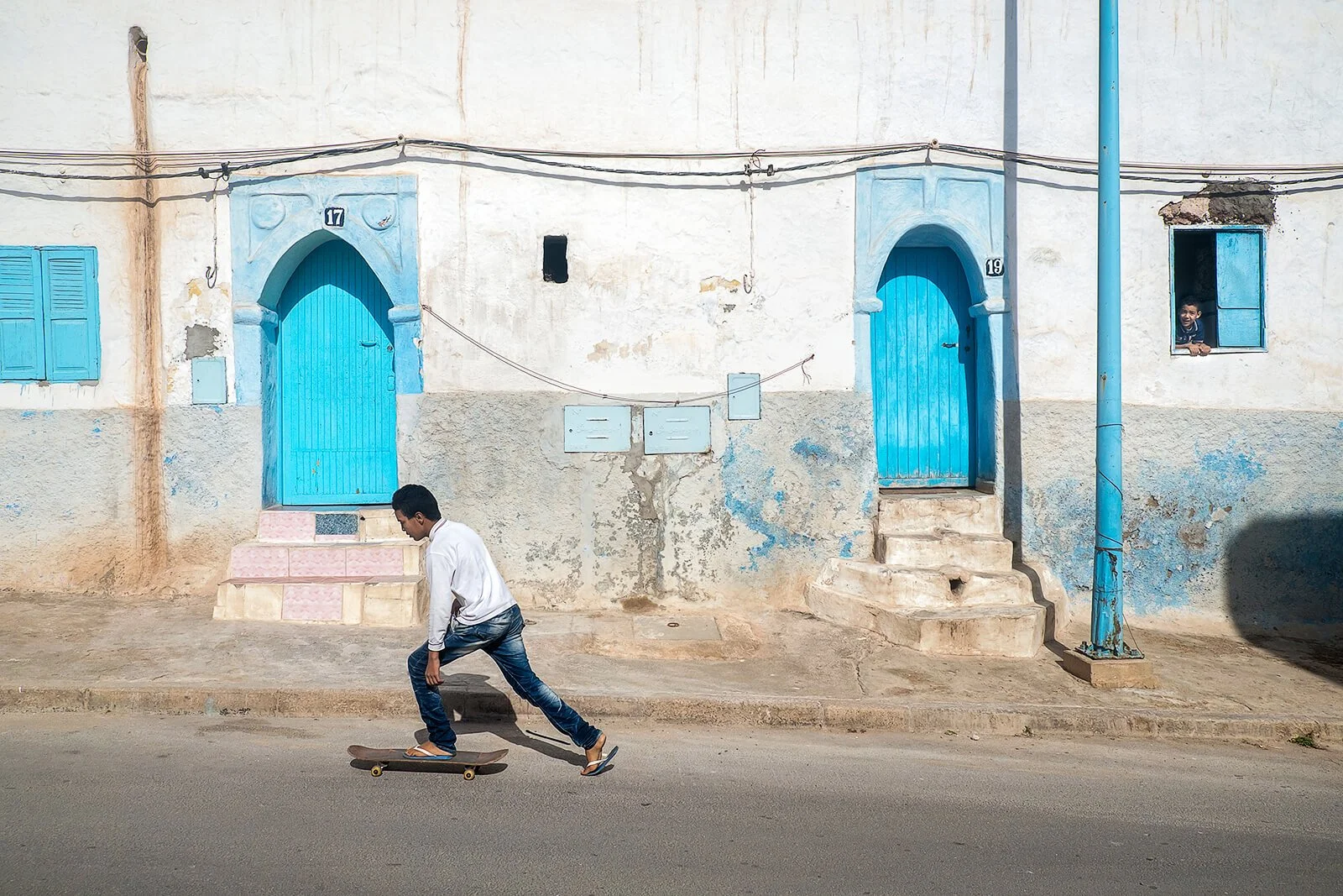 Sidi Ifni, Maroc — un skateur remonte la rue sous le regard d’un jeune garçon à la fenêtre.