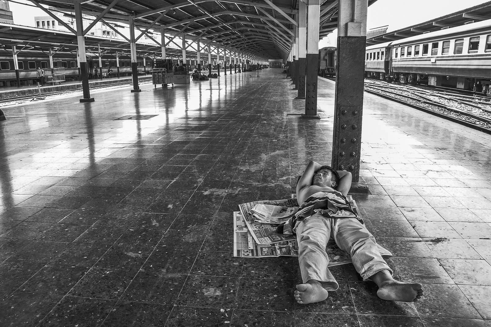 Street photography noir et blanc : un homme dort sur des journaux étalés sur le sol de marbre de la gare de Bangkok. Traitement dense et contrasté.