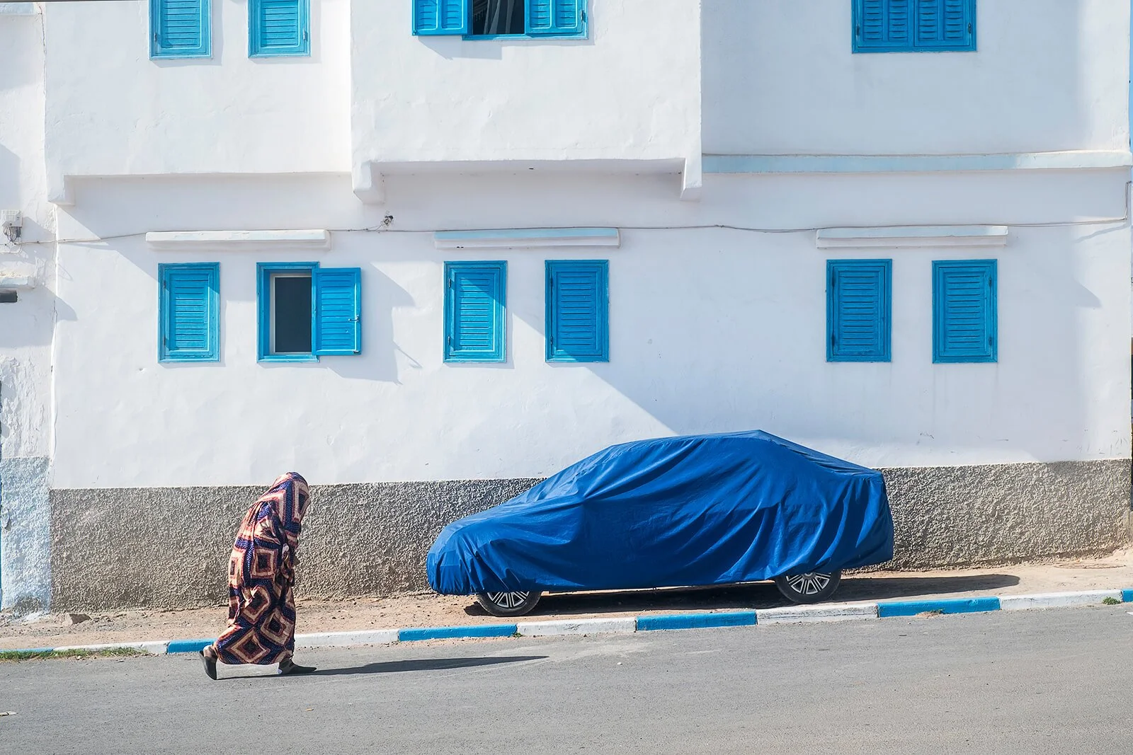 Sidi Ifni, Maroc — une silhouette enveloppée dans une couverture passe devant une voiture protégée par une bâche bleue.