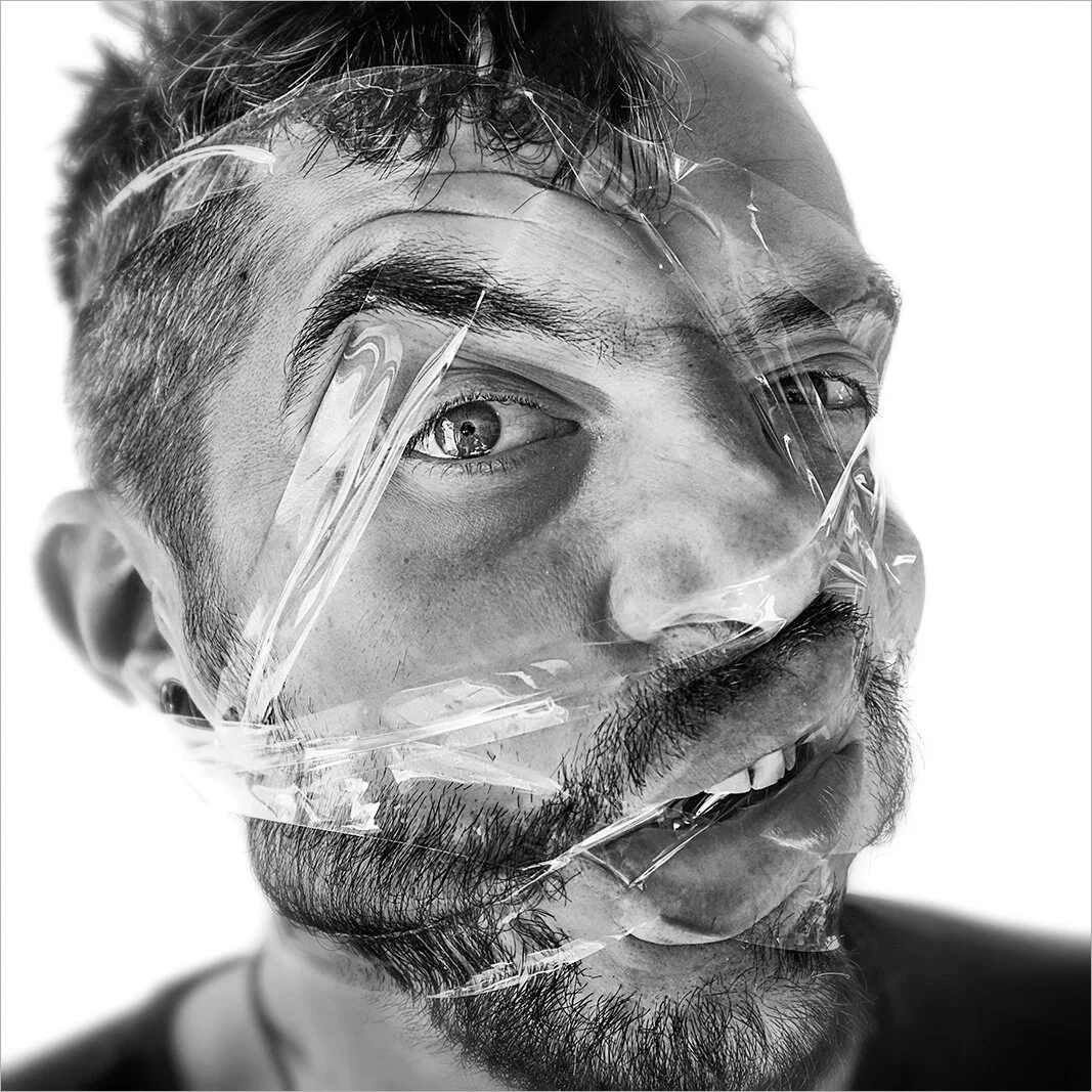 Portrait humoristique noir et blanc d'un homme aux yeux écarquillés, luttant contre un ruban adhésif qui lui compresse le nez et les joues. Une grimace expressive capturée par Hugues Hardy.