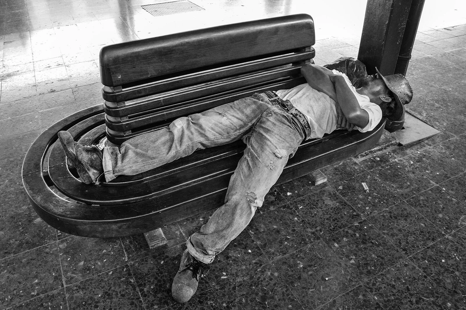 Portrait environnemental noir et blanc : un homme au chapeau fait la sieste sur un banc en bois de la gare Hua Lamphong. Photographie de rue, Thaïlande.