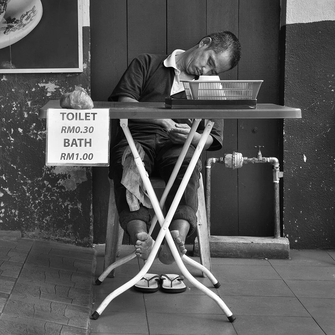 Portrait sur le vif en noir et blanc à Melaka en Malaisie : un employé de sanitaires s'est assoupi sur sa chaise. Photo Hugues Hardy.