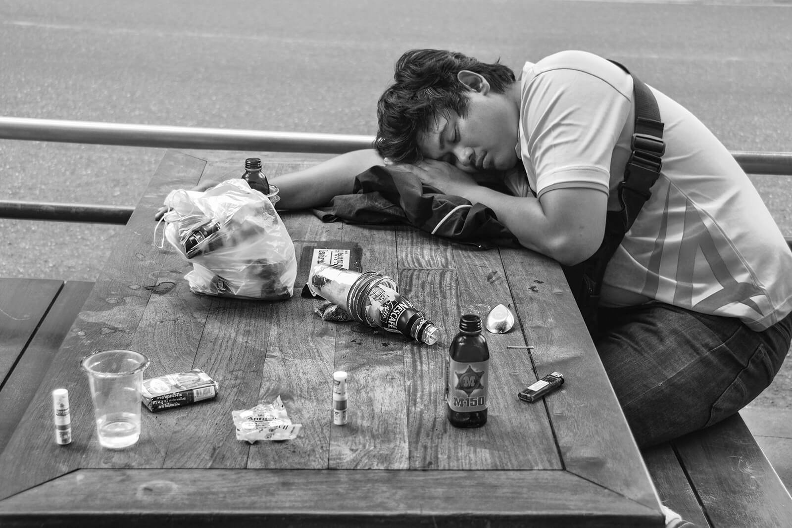 Reportage photo noir et blanc en Thaïlande : un jeune homme endormi sur une table jonchée de bouteilles sur un trottoir de Bangkok. Traitement dense.