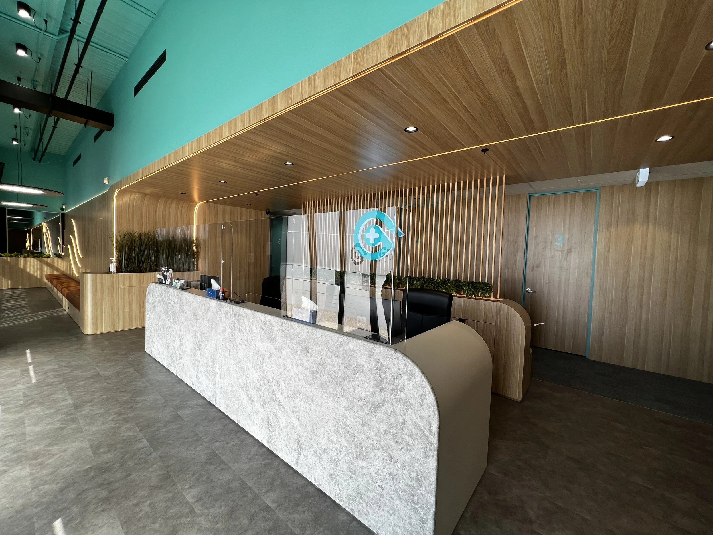 Modern reception area with a white stone reception desk, a glass partition, and wood paneling with creative lighting. There are plants on the side, and two doorways in the background, numbered 3 and 6.